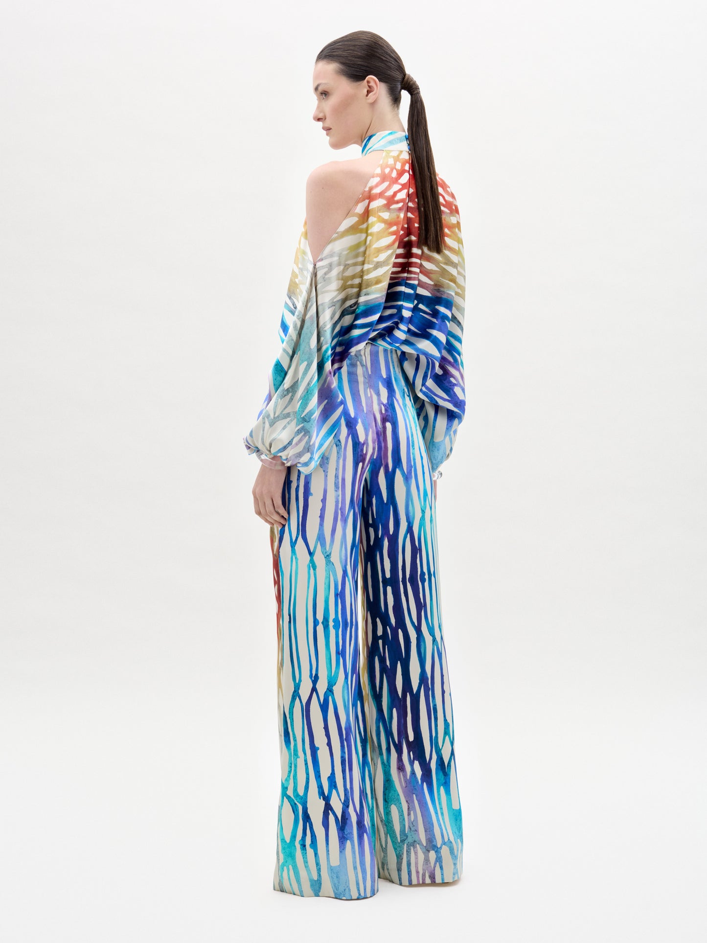 Una persona con una coleta baja, de espaldas, lleva el Andie Pant: un mono de cuello halter con pantalones de cintura alta y un colorido estampado abstracto de rayas rojas, azules, amarillas y moradas. El fondo es blanco liso.