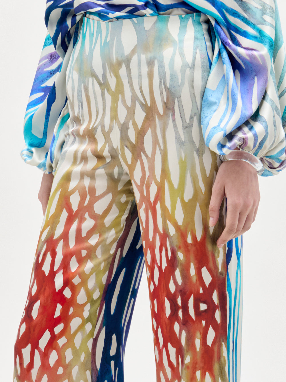 Una persona lleva el pantalón Andie, un modelo de cintura alta de colores vivos con un estampado abstracto rojo, amarillo y azul, y una camiseta de manga larga a juego con dibujos azules y blancos; sólo se ven la parte inferior del torso y los brazos.