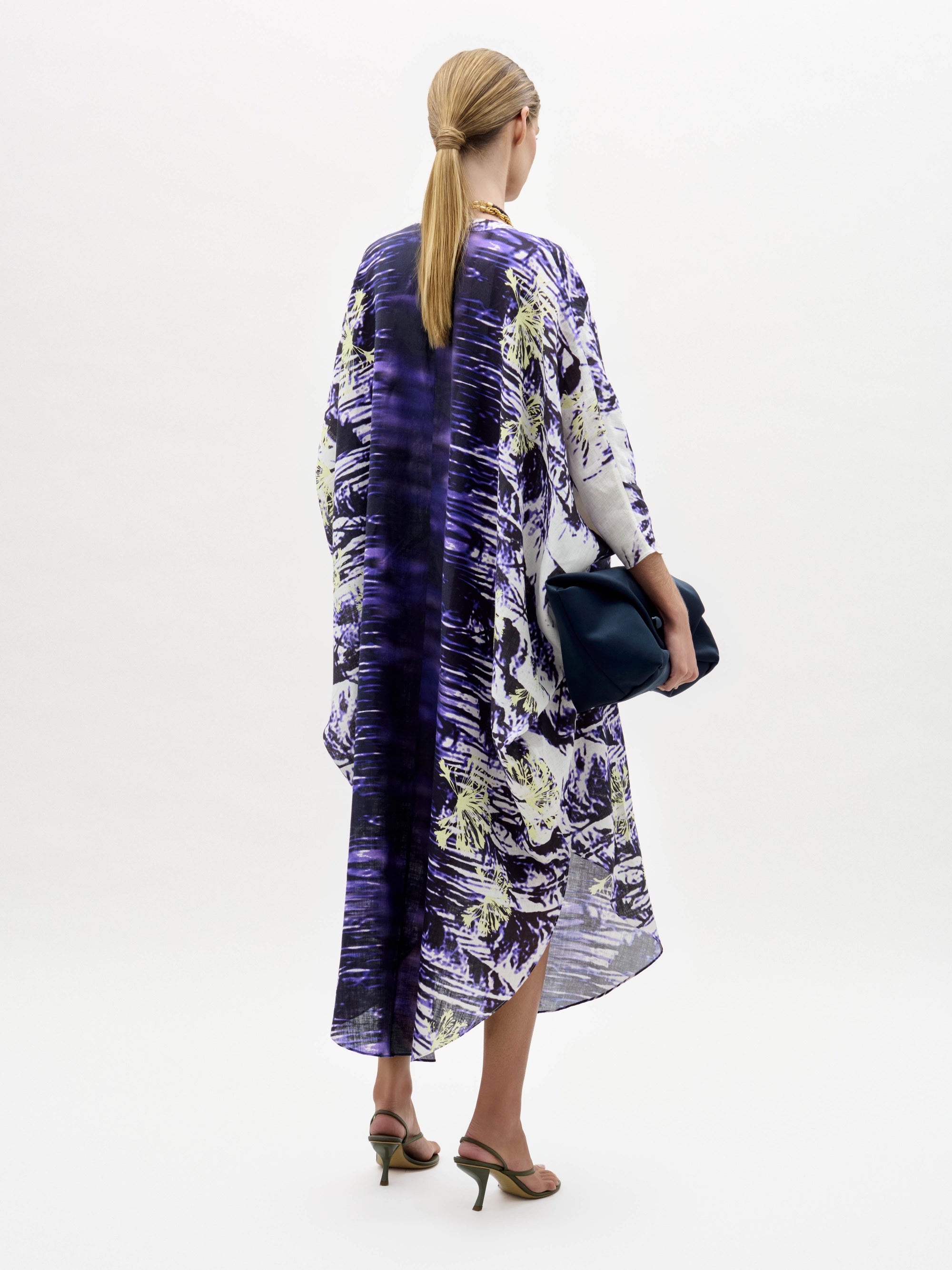 Una persona, de espaldas, lleva el vestido Ilse, suelto y hasta la rodilla, con un estampado abstracto en morado, blanco y amarillo. Lleva un bolso de mano azul oscuro, el pelo castaño claro recogido y sandalias de tacón.