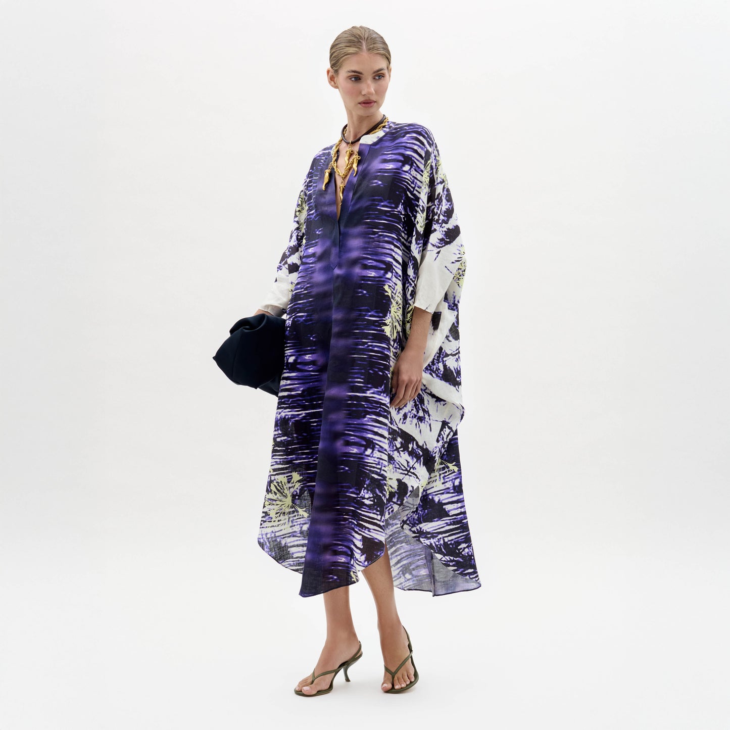 Una mujer, sobre un fondo blanco liso, lleva el vestido Ilse, un caftán suelto con estampado morado y blanco. Lo combina con sandalias de tacón y un bolso oscuro. Lleva el pelo recogido y su expresión es neutra.