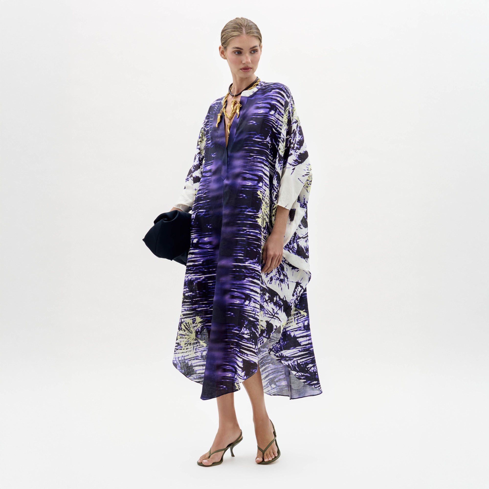 Una mujer, sobre un fondo blanco liso, lleva el vestido Ilse, un caftán suelto con estampado morado y blanco. Lo combina con sandalias de tacón y un bolso oscuro. Lleva el pelo recogido y su expresión es neutra.