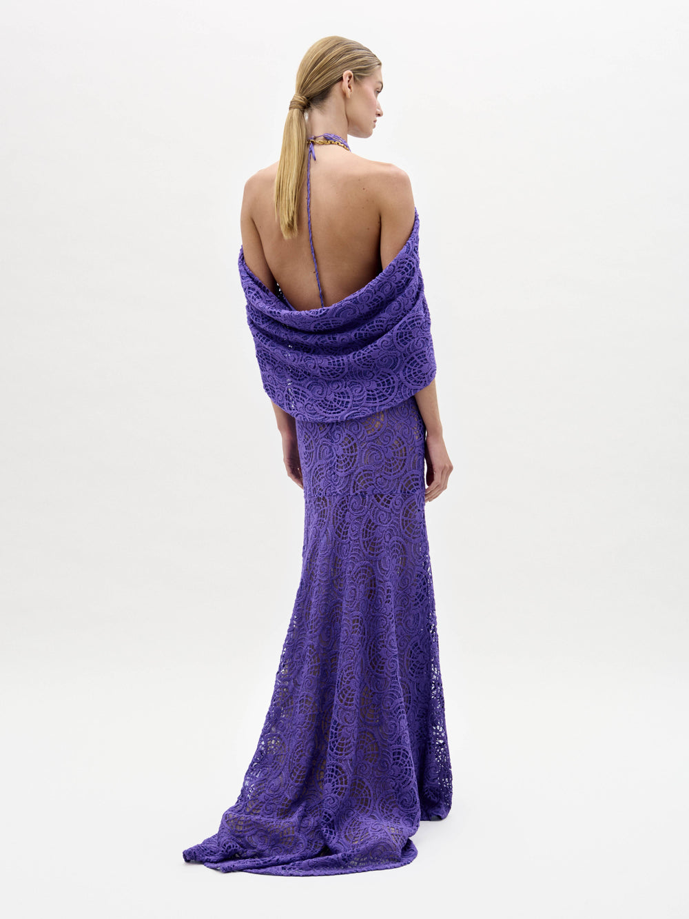 Una persona de pie sobre un fondo liso lleva el vestido Salvina, un vestido de encaje morado hasta el suelo, sin espalda, con cuello halter y detalle de chal drapeado. Lleva el pelo rubio, largo y liso, recogido en una coleta baja.