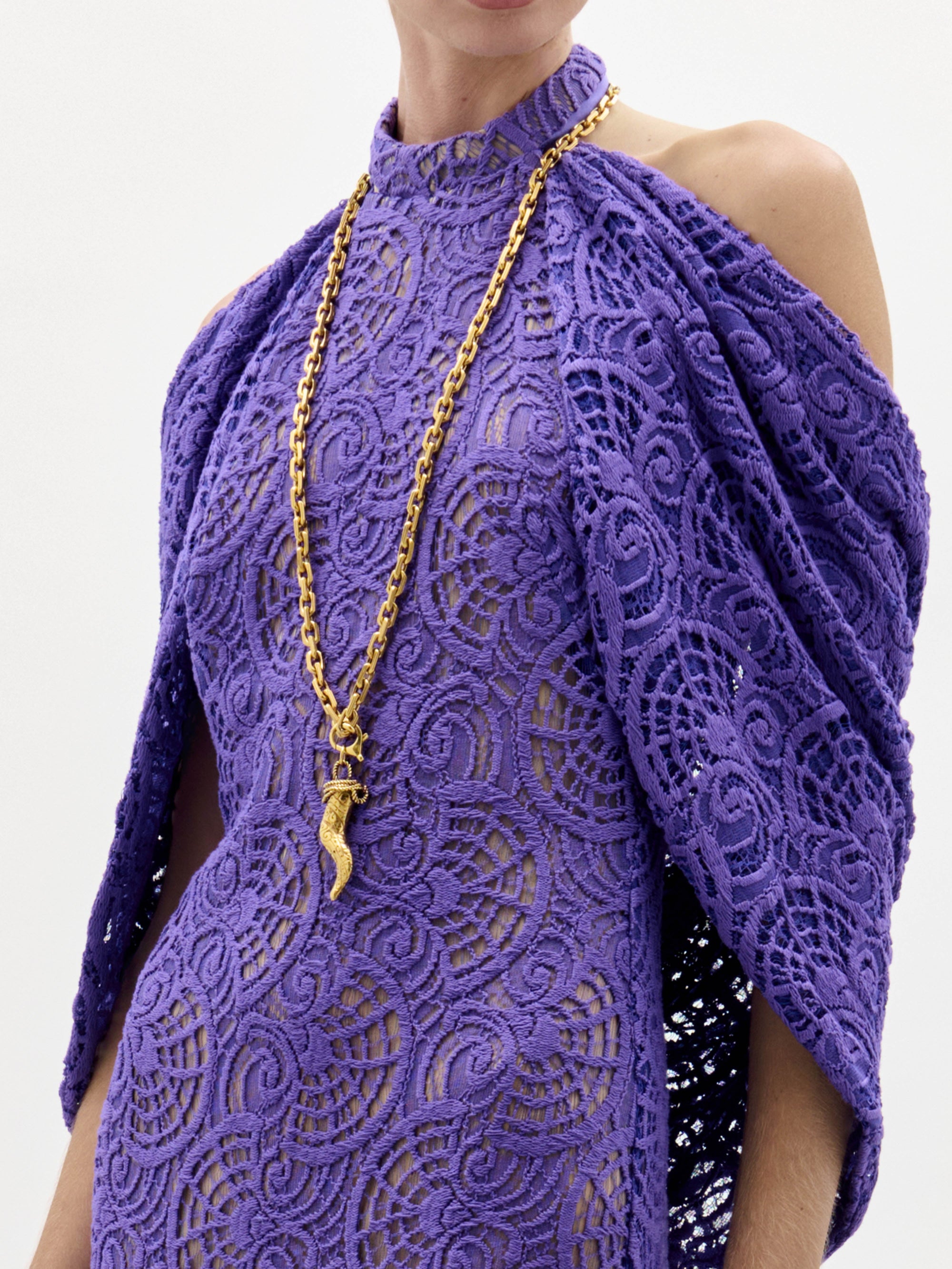 Una persona lleva el vestido Salvina -un vestido de encaje morado de cuello alto con un detallado diseño en espiral y un chal adosado- combinado con un collar de cadena dorada con un gran colgante.