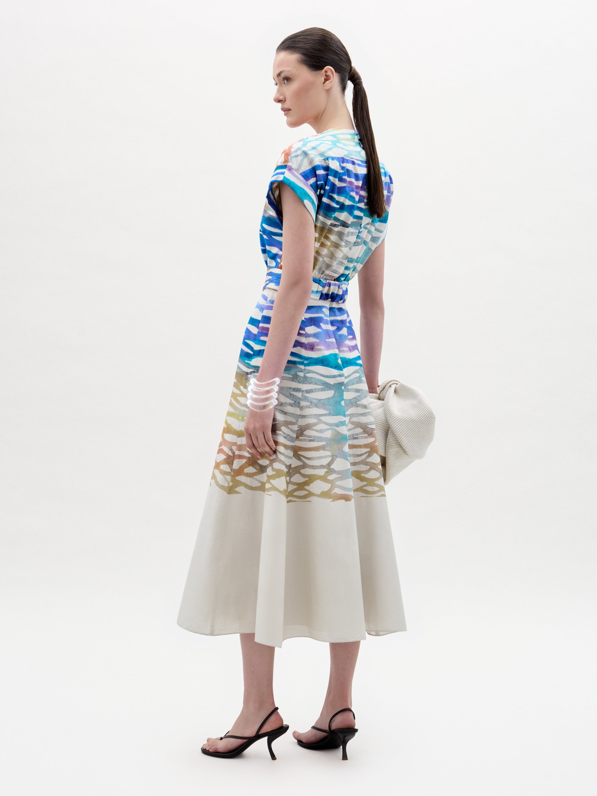 Una mujer luce de perfil el vestido Sakura, un colorido vestido midi estampado con falda acampanada, combinado con tacones de tiras negros, brazaletes blancos y un bolso de mano blanco con textura; un inconfundible estilo colombiano sobre un fondo blanco liso.
