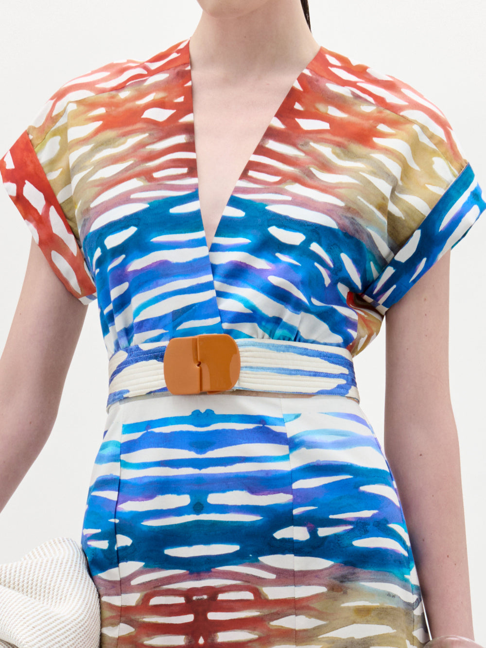 Una persona lleva el vestido Sakura, un midi con cuello en V y rayas horizontales abstractas de colores rojo, azul, dorado y blanco, ceñido a la cintura por un cinturón beige con una gran hebilla marrón. Hecho en Colombia.