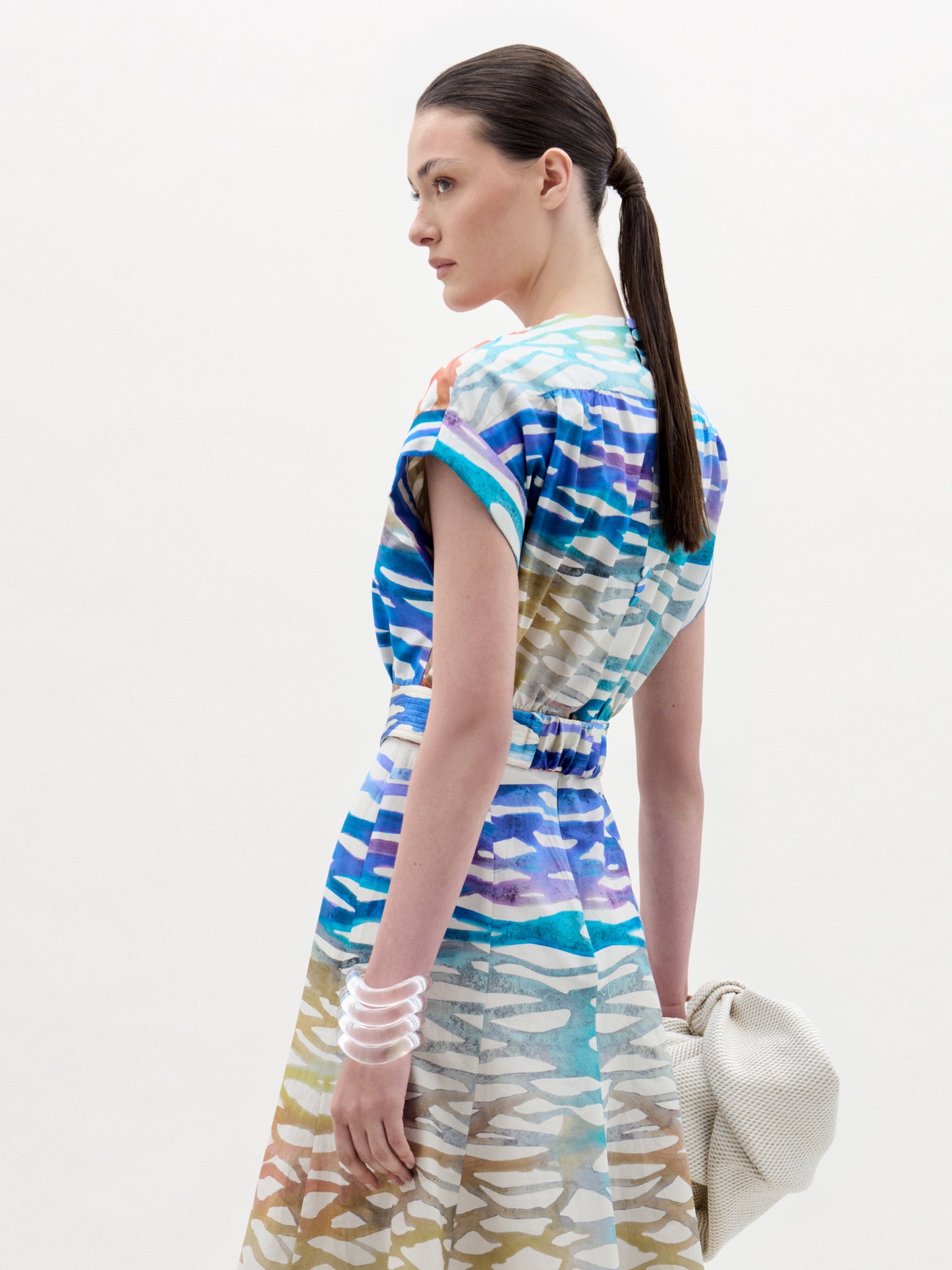Una mujer con el pelo castaño liso recogido en una coleta baja lleva la blusa Bara, con un estampado abstracto azul, blanco y tostado. Sostiene un bolso de color claro con textura, lleva varias pulseras transparentes y mira ligeramente hacia un lado.