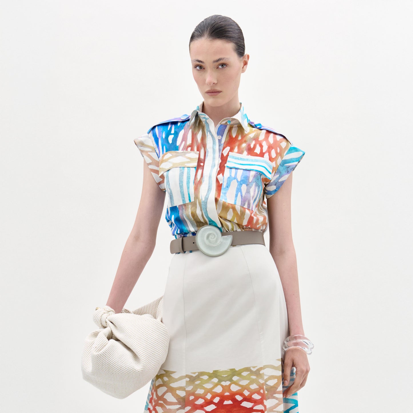 Una mujer, sobre fondo blanco, lleva la blusa Bara, un top manga corta de algodón de colores fabricado en Colombia, metido dentro de una falda crema con dobladillos a juego. Lleva un bolso beige con textura y un cinturón ancho con hebilla circular.