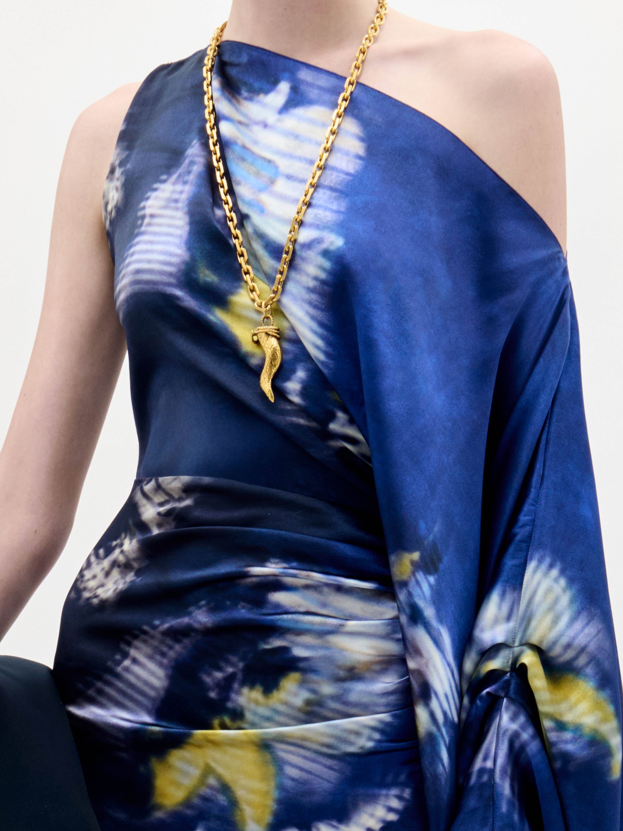 Una persona lleva el vestido Dotty, una prenda de un solo hombro estampada en azul y blanco, combinada con un collar de cadena dorada con un colgante en forma de serpiente, sobre un fondo liso de color claro.