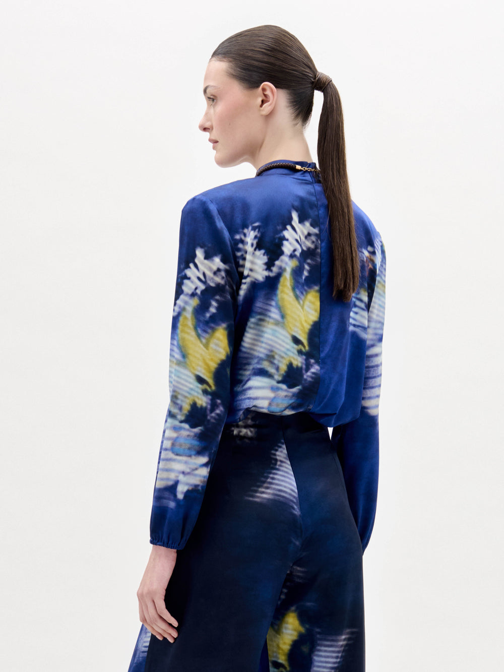 Una persona con el pelo castaño liso recogido en una coleta baja aparece de espaldas, con la blusa Lane -una blusa azul con motivos florales y abstractos en amarillo, blanco y azul oscuro- sobre un fondo claro liso.