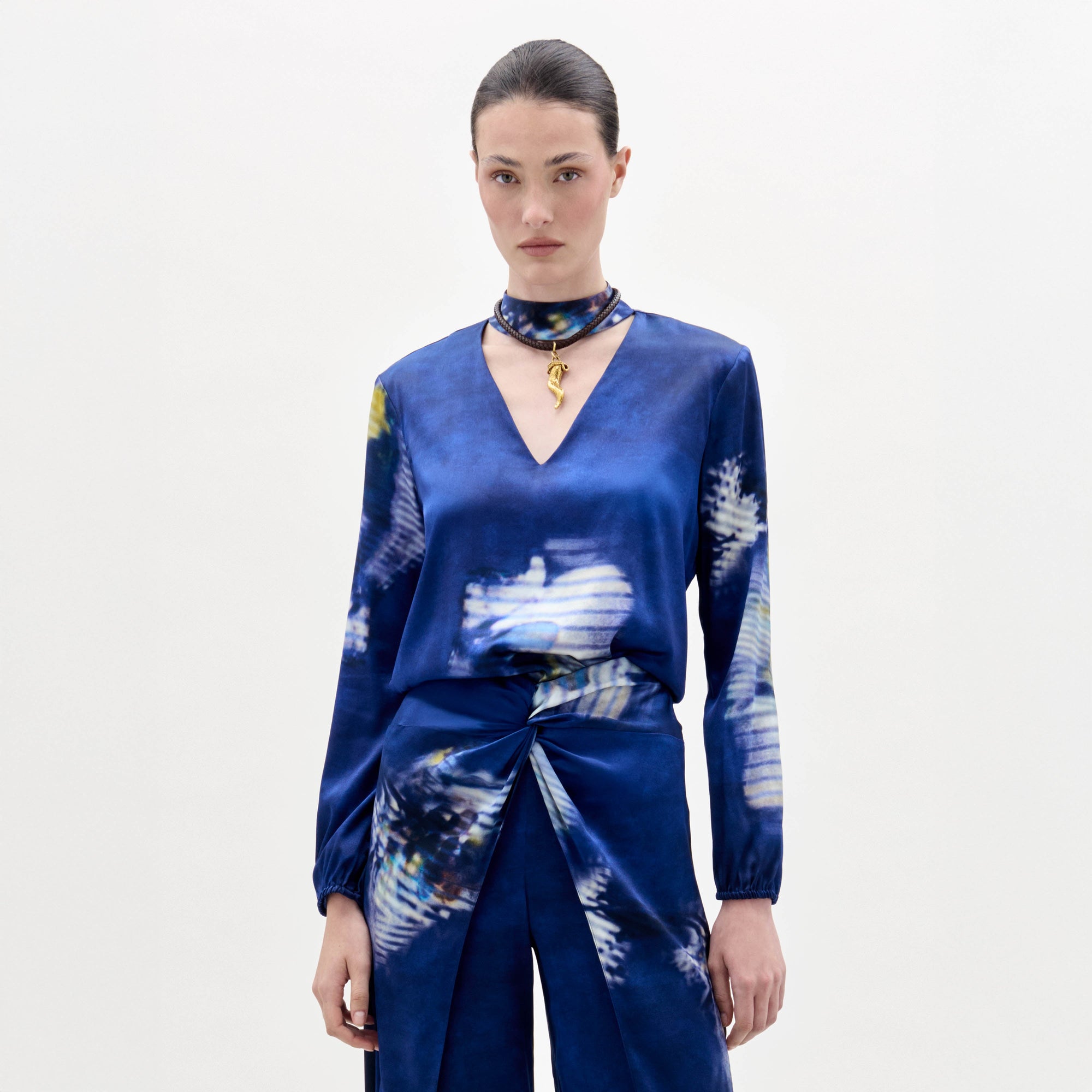 Una mujer con el pelo peinado hacia atrás lleva la blusa Lane -una blusa azul estampada de manga larga y cuello en V- combinada con pantalones a juego y una gargantilla con colgante dorado sobre un fondo blanco liso.