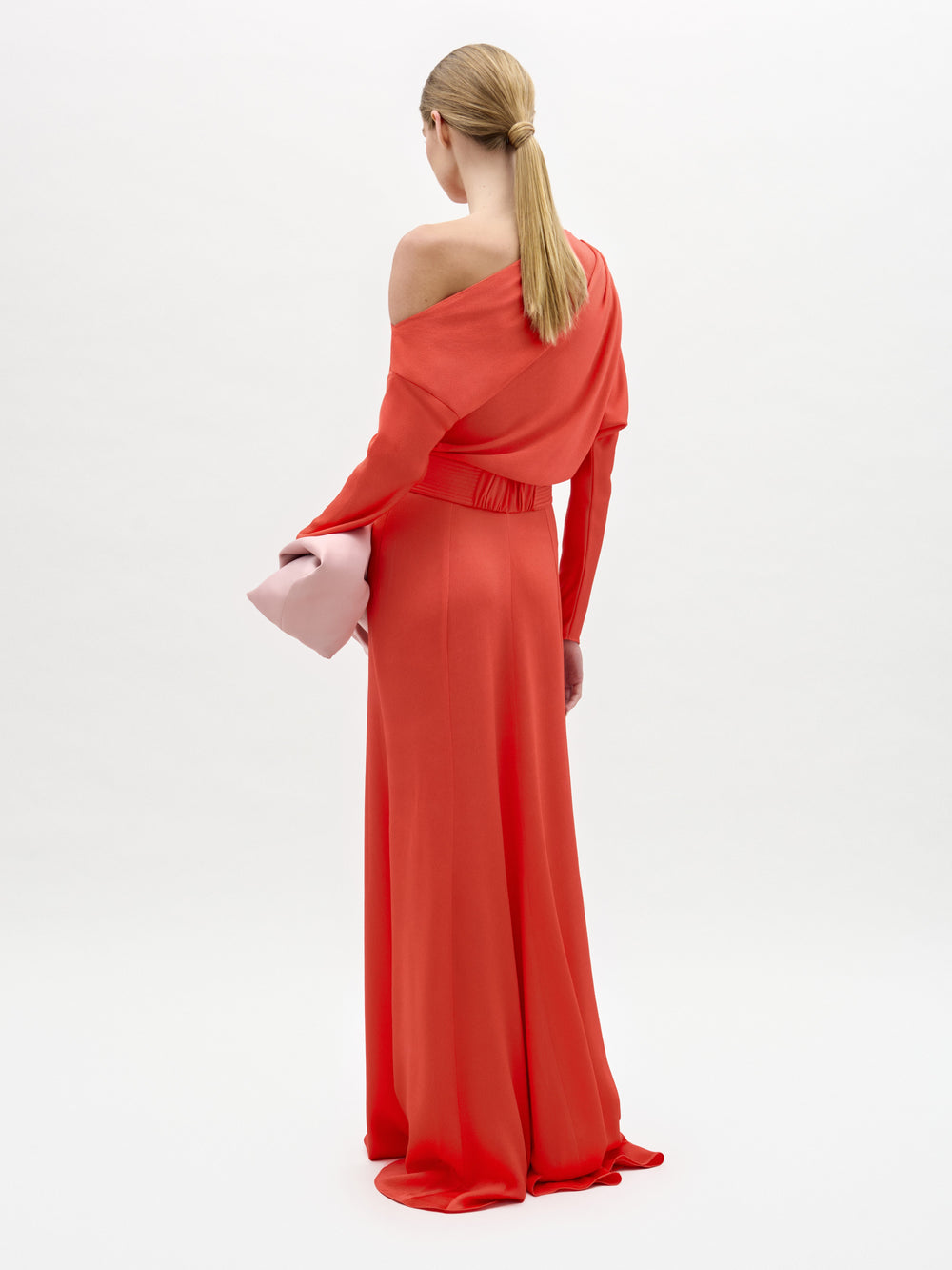 Una mujer con el pelo rubio recogido en una coleta baja lleva el vestido rojo brillante Noelia Dress, de una sola manga, y un bolso de mano rosa claro. Está de pie sobre un fondo blanco liso, de espaldas a la cámara.