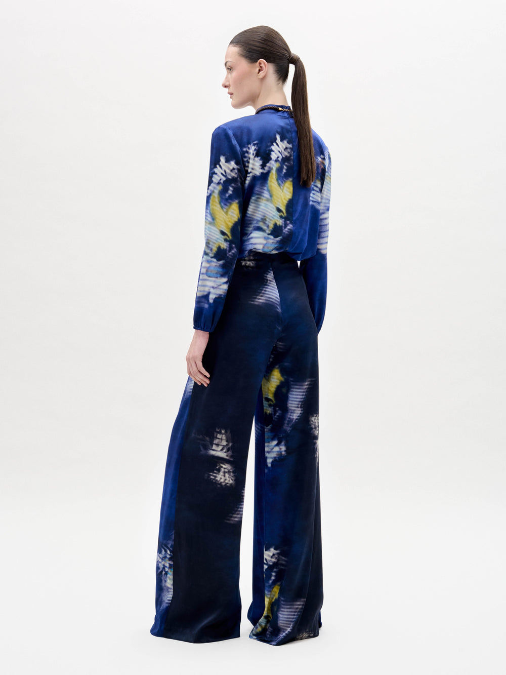 Una persona de pie, de espaldas, lleva el Pantalón Canturipe en azul oscuro con una silueta de pierna ancha a juego y un top de manga larga adornado con motivos florales abstractos en blanco, amarillo y azul pálido. Lleva el pelo recogido en una coleta baja.