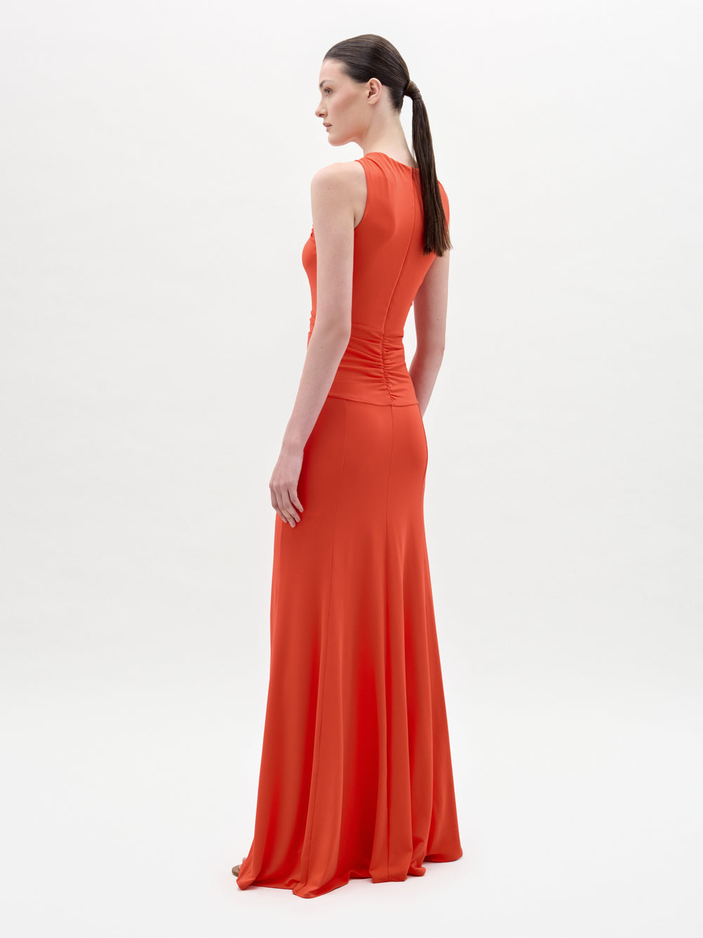 Una mujer, de perfil, lleva el vestido Naga, un maxi naranja sin mangas, largo hasta el suelo, con cuerpo ajustado y falda vaporosa. Lleva el pelo recogido en una coleta baja sobre un fondo liso y claro.