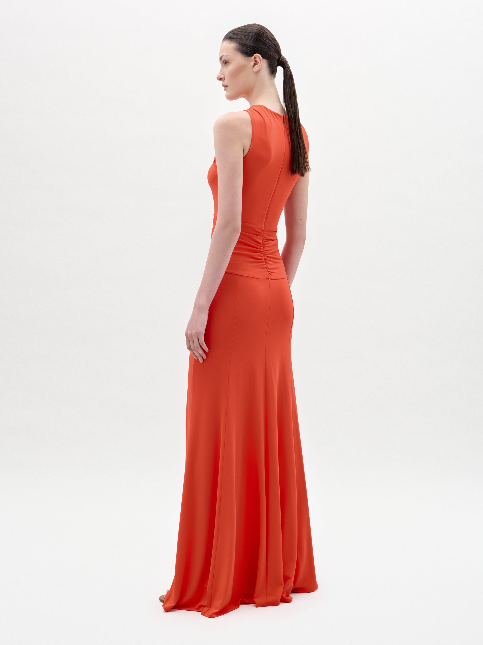 Una mujer, de perfil, lleva el vestido Naga, un maxi naranja sin mangas, largo hasta el suelo, con cuerpo ajustado y falda vaporosa. Lleva el pelo recogido en una coleta baja sobre un fondo liso y claro.