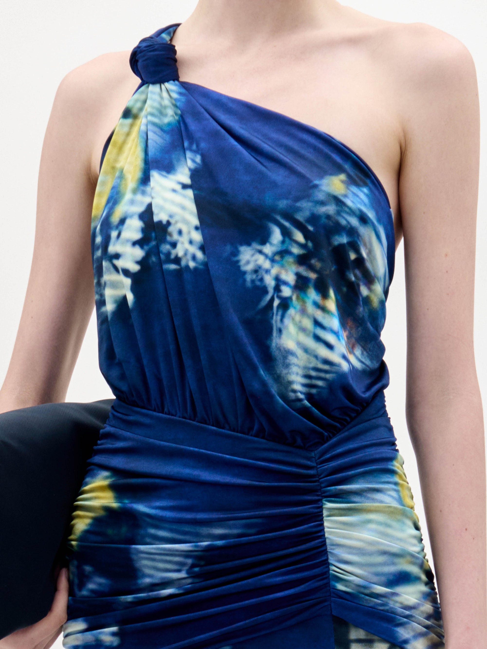 Una persona lleva el vestido Antara, un modelo azul de un solo hombro con un nudo fruncido en el hombro, cintura fruncida y un estampado tie-dye en tonos azules, blancos y amarillos.