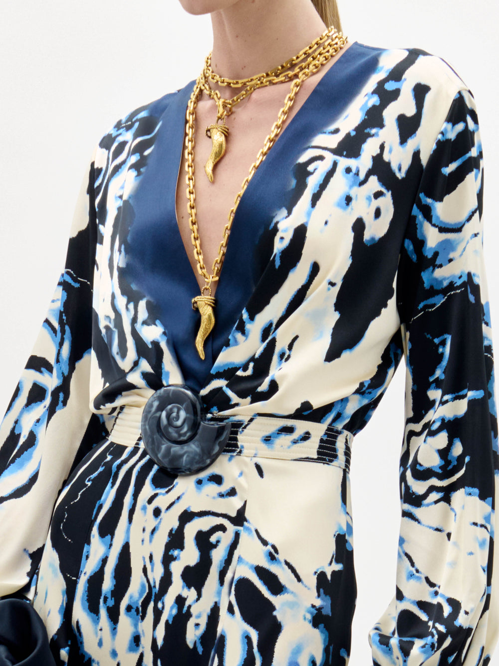 Una persona lleva el vestido Remy -un vestido de manga larga, estampado en azul y blanco, con escote profundo en V, cinturón ancho con hebilla redonda- combinado con dos collares de cadena de oro con colgantes en forma de cuerno.