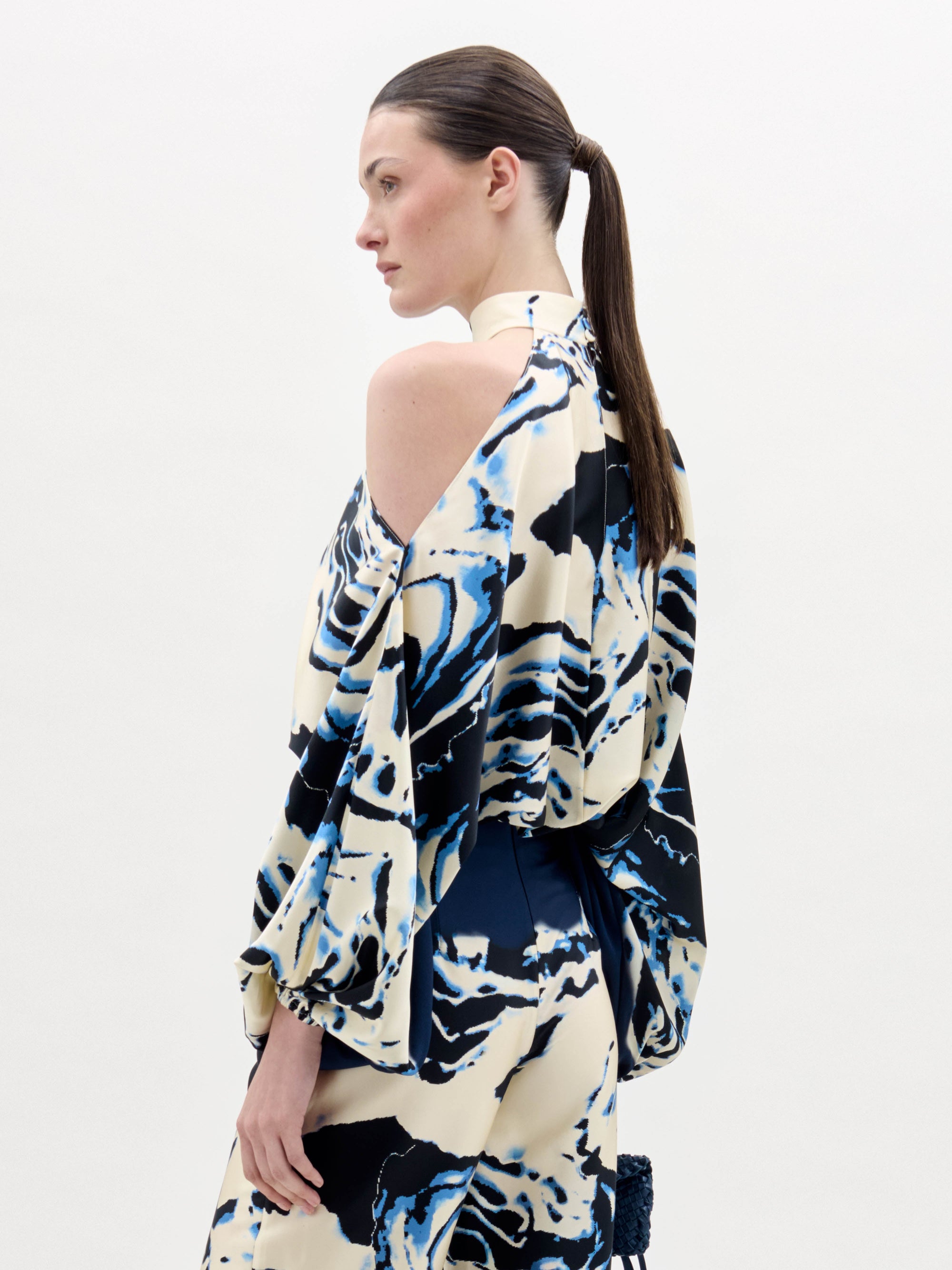 Una persona con el pelo largo y castaño recogido en una coleta baja lleva la blusa Safira, un top de cuello halter con motivos abstractos en azul, negro y crema y un hombro al descubierto. El fondo es blanco liso.