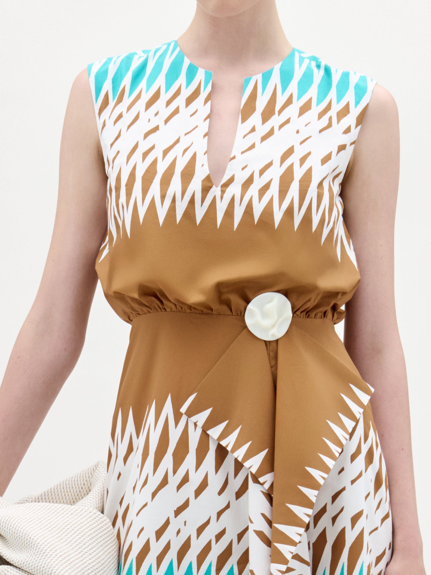 Una persona lleva el vestido Kendall, un vestido midi marrón sin mangas con motivos geométricos en blanco y turquesa, escote con abertura delantera, silueta fit & flare, cintura fruncida con un gran botón blanco. Sostienen tela italiana de color claro.
