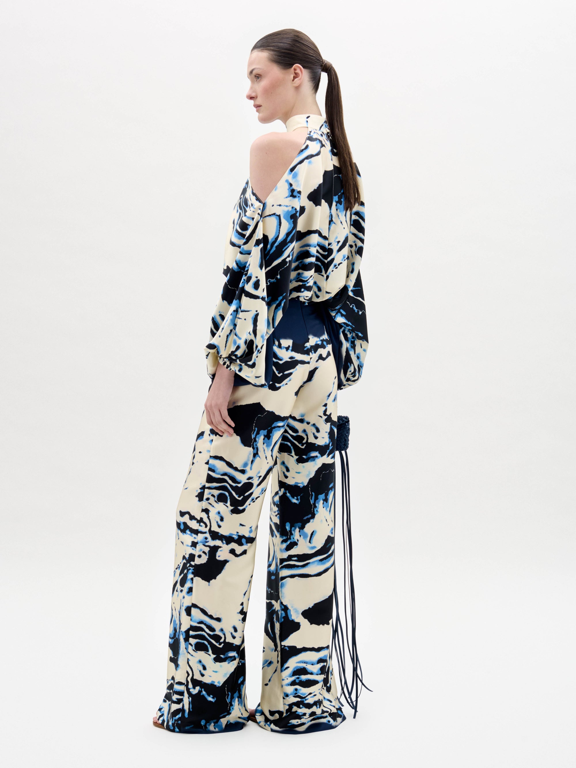 Una persona de pie, de lado, sobre un fondo claro y liso, lleva el pantalón Como -de pierna ancha y holgada, con un estampado abstracto en colores crema, azul marino y negro- combinado con un top a juego de hombros fríos. Lleva el pelo recogido en una coleta baja.