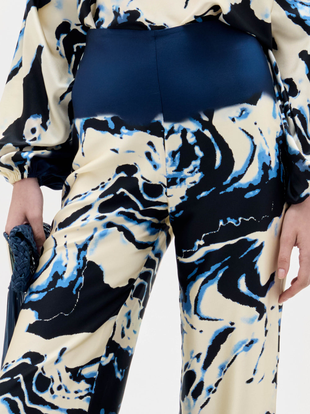 Una persona lleva el pantalón Como -de talle alto y estampado jaspeado en azul, negro y crema- y un top de manga larga a juego. Una mano sujeta un bolso de mano azul marino con textura. La imagen muestra desde los hombros hasta medio muslo.