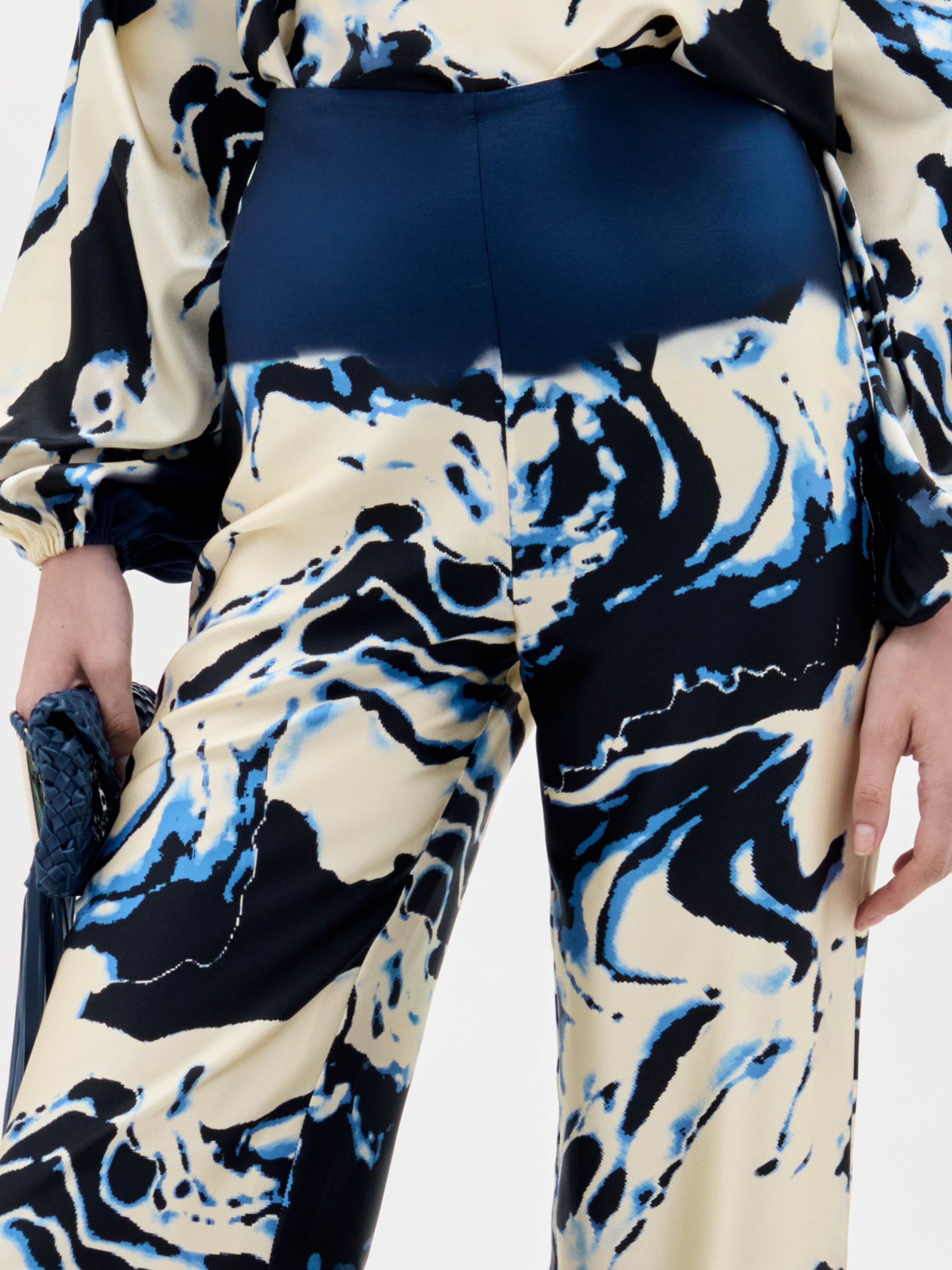 Una persona lleva el pantalón Como -de talle alto y estampado jaspeado en azul, negro y crema- y un top de manga larga a juego. Una mano sujeta un bolso de mano azul marino con textura. La imagen muestra desde los hombros hasta medio muslo.