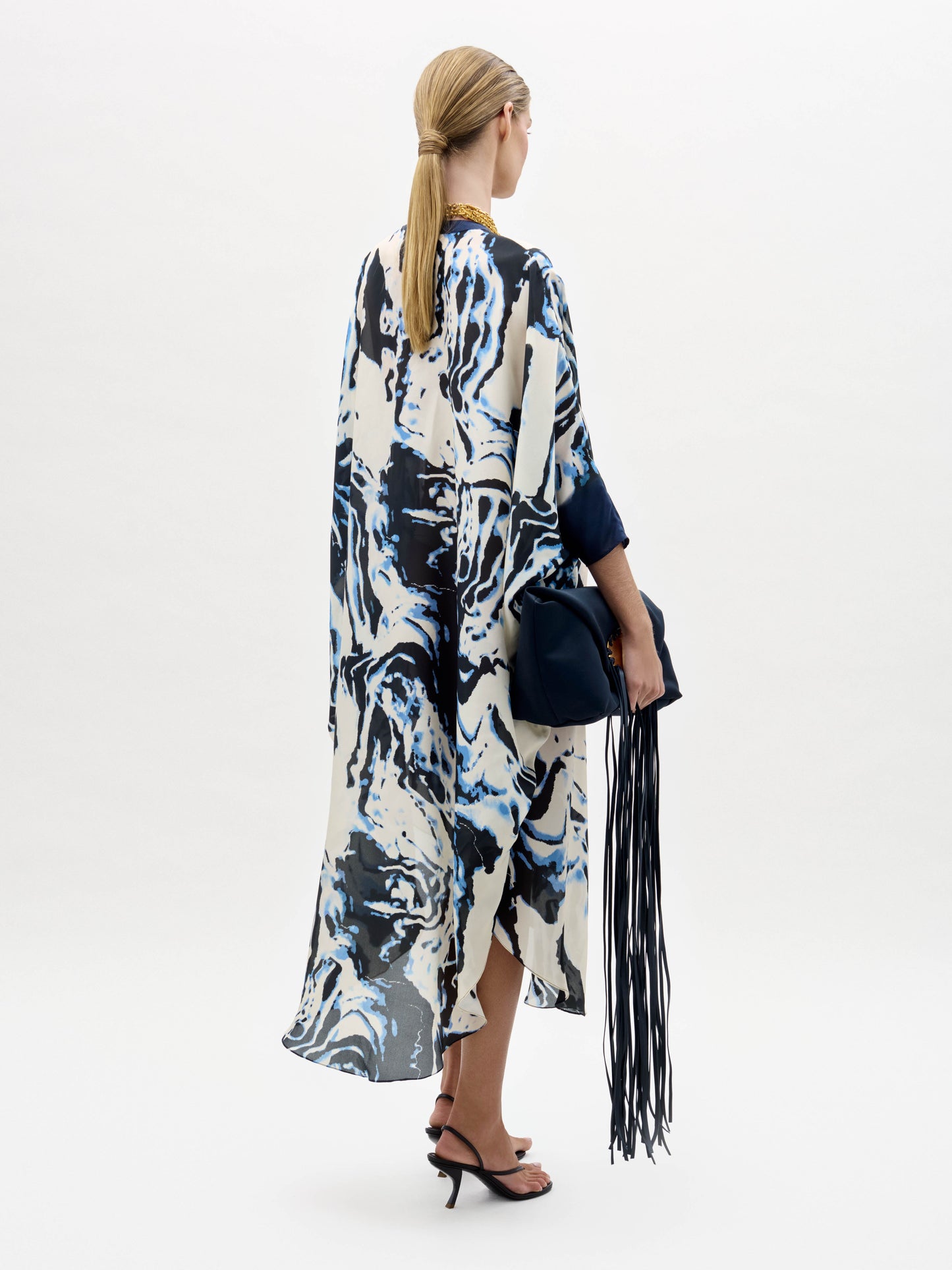 Una persona con el pelo rubio recogido en una coleta baja lleva la túnica Elea -un vestido suelto estampado en azul y blanco- con sandalias de tacón negras y sostiene un bolso azul marino con flecos sobre un fondo blanco liso.