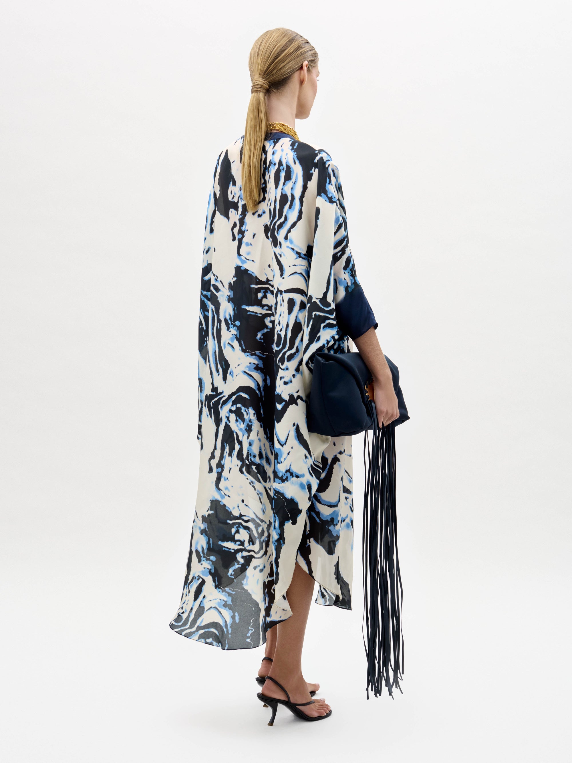 Una persona con el pelo rubio recogido en una coleta baja lleva la túnica Elea -un vestido suelto estampado en azul y blanco- con sandalias de tacón negras y sostiene un bolso azul marino con flecos sobre un fondo blanco liso.