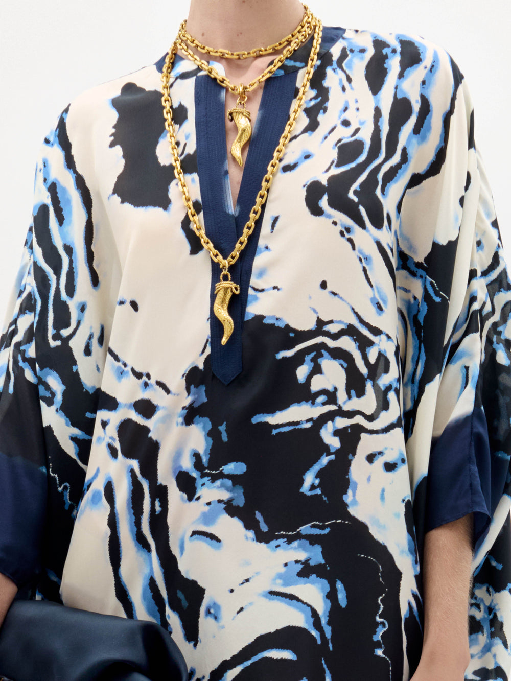 Lleva la túnica Elea con motivos azul marino, crema y azul claro, y dos collares de cadena dorada con colgantes abstractos. No se ve la cabeza de la persona.