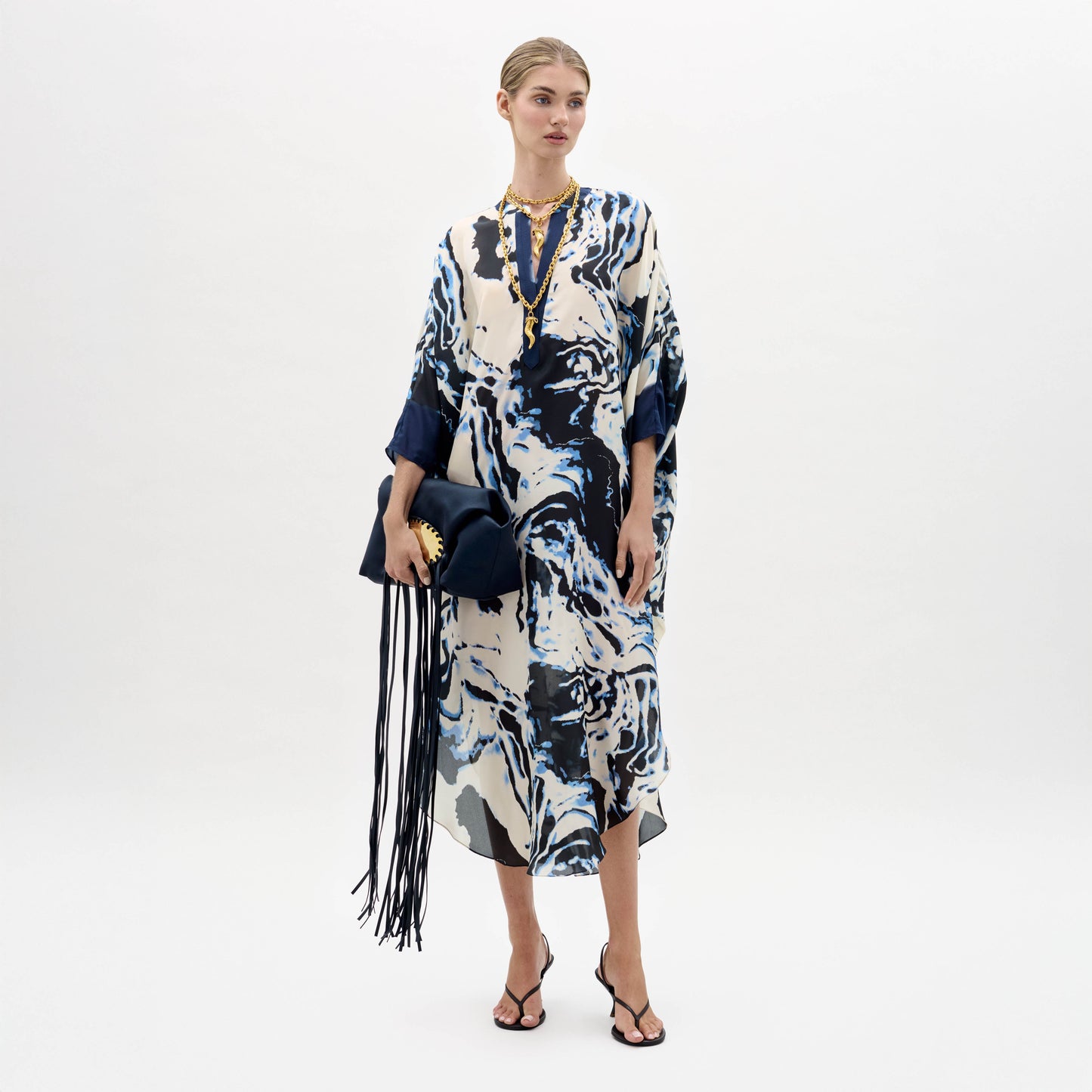 Sobre un fondo blanco, una mujer lleva la túnica Elea -un vestido suelto con motivos marmóreos en azul y blanco- combinada con sandalias negras, un collar dorado y un bolso azul marino con flecos.