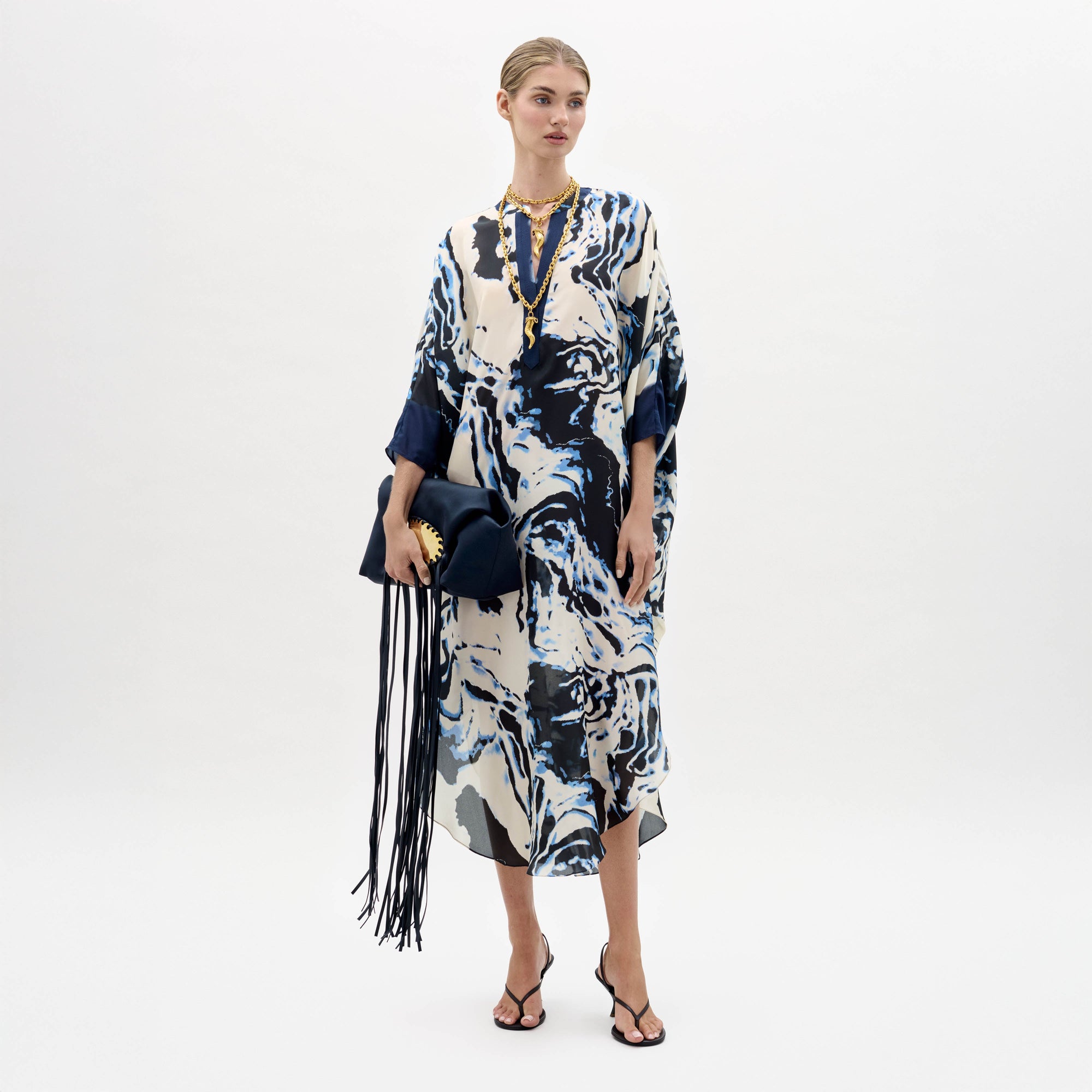 Sobre un fondo blanco, una mujer lleva la túnica Elea -un vestido suelto con motivos marmóreos en azul y blanco- combinada con sandalias negras, un collar dorado y un bolso azul marino con flecos.