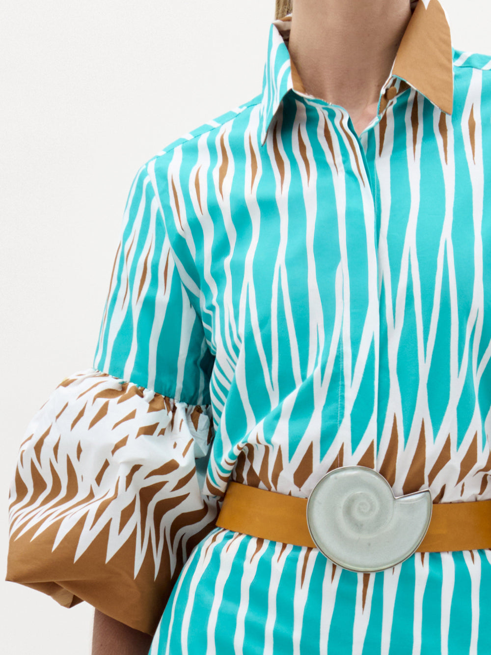 Una persona lleva la blusa Helina, con estampados en zigzag azul y marrón, cuello marrón, mangas abullonadas y cinturón ancho con hebilla de concha grande: un look elegante que muestra la calidad hecho en Colombia.