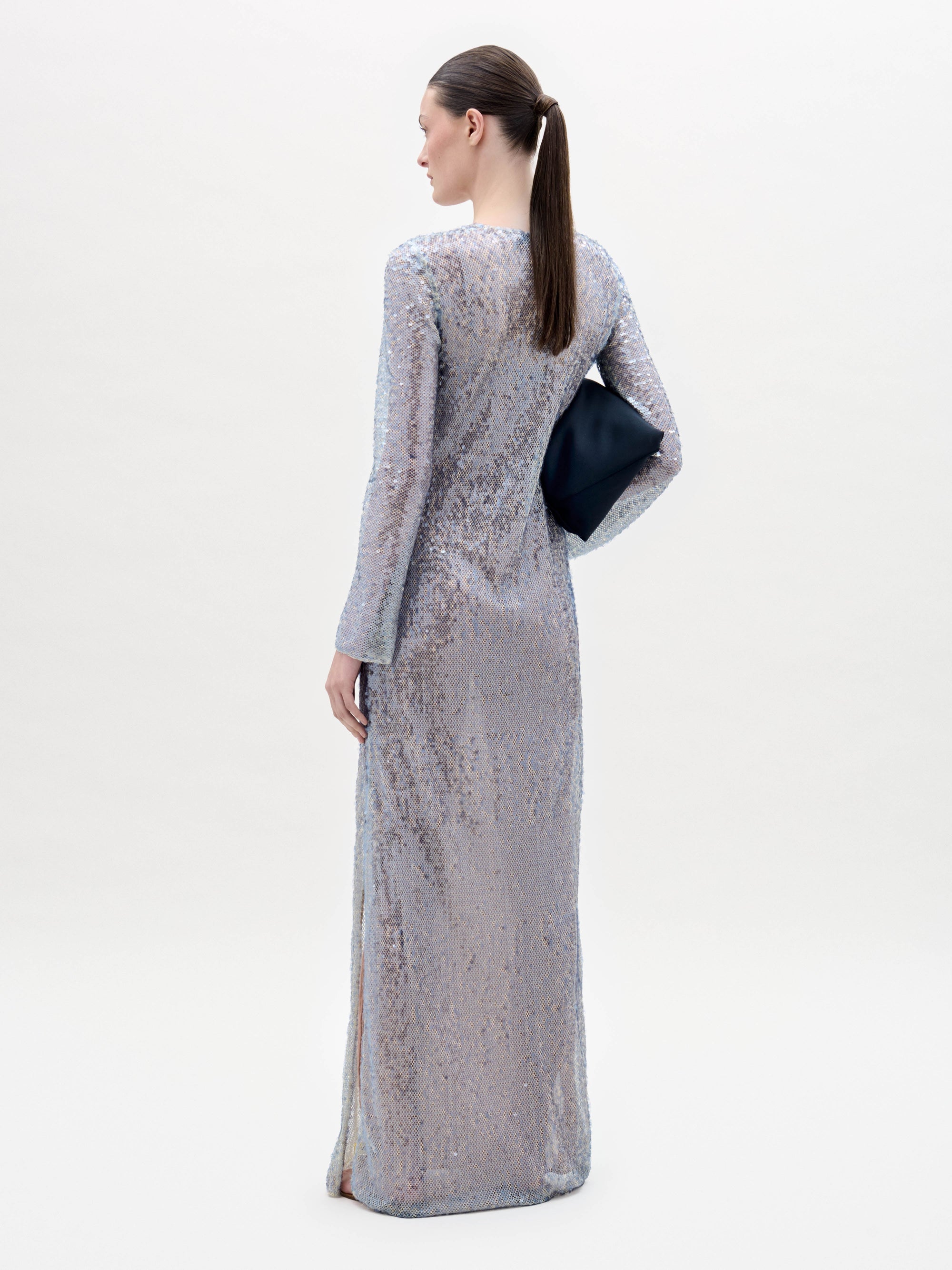 Una persona con el pelo castaño liso recogido en una coleta baja luce el vestido Nico -un vestido largo de lentejuelas azul claro- con un clutch negro en la mano y de pie sobre un fondo claro y liso, de espaldas a la cámara.