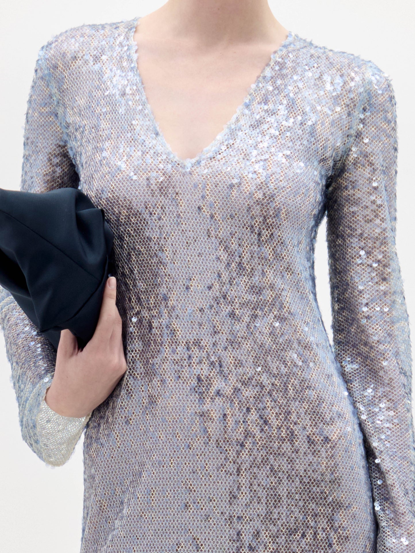 Con el vestido Nico -de manga larga y cuello en V de lentejuelas plateadas-, una persona lleva un bolso negro doblado en la mano izquierda sobre un fondo claro liso.