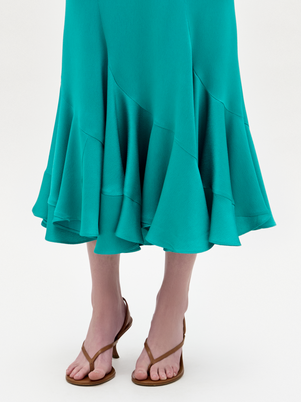 Una persona con la falda Marlene en color verde azulado, con tejido italiano y volantes, la combina con sandalias marrones de puntera abierta sobre un fondo claro liso.
