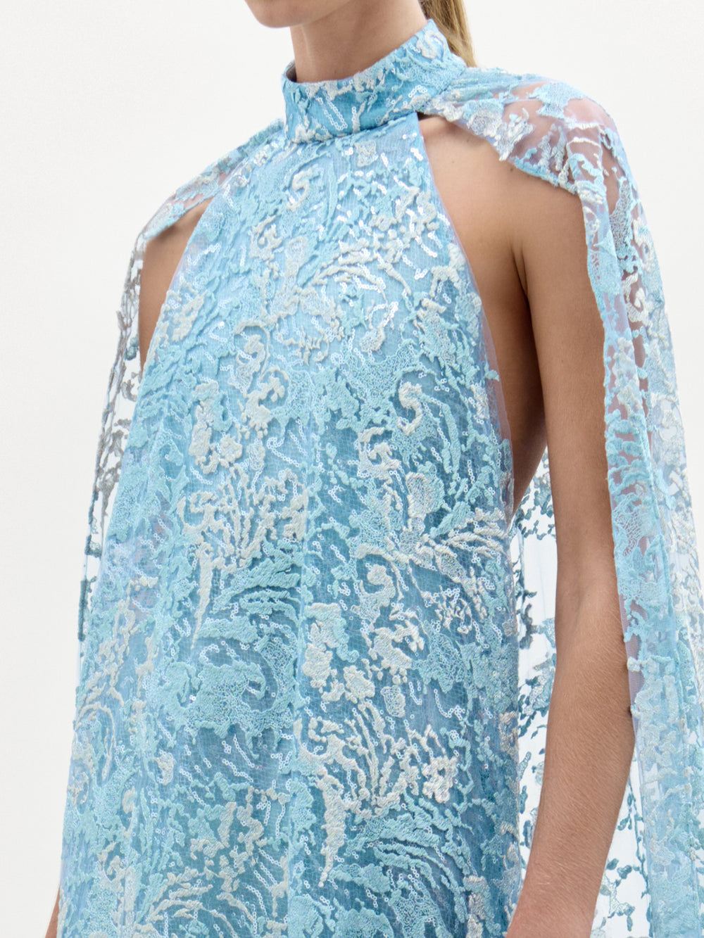 Una persona lleva el Vestido Carol, un vestido de columna sin mangas y cuello alto hecho a mano en Colombia a partir de encaje floral azul claro con hombros en forma de capa y detalles parcialmente transparentes. El rostro de la portadora no es visible.