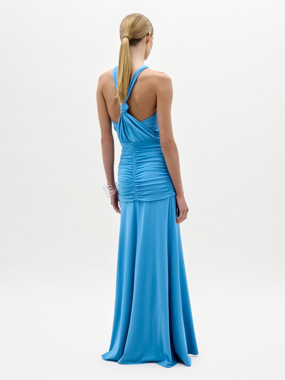 Una mujer con el pelo rubio liso recogido en una coleta baja mira de frente y lleva el vestido Jedda, un maxi largo de tejido italiano azul brillante con frunces, espalda abierta y un brazalete plateado. Fabricado en Colombia.