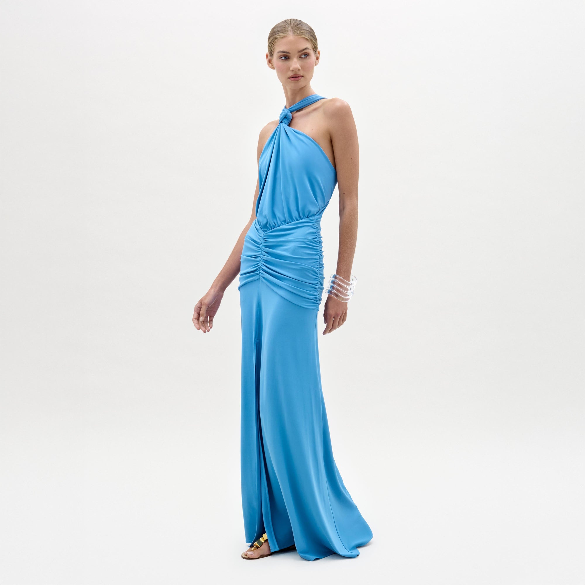 Una mujer posa sobre un fondo blanco liso con el vestido Jedda, un maxi largo de color azul brillante con anudado delantero y cintura fruncida, confeccionado en tejido italiano. Lleva el pelo recogido y accesorios como sandalias y brazaletes transparentes.