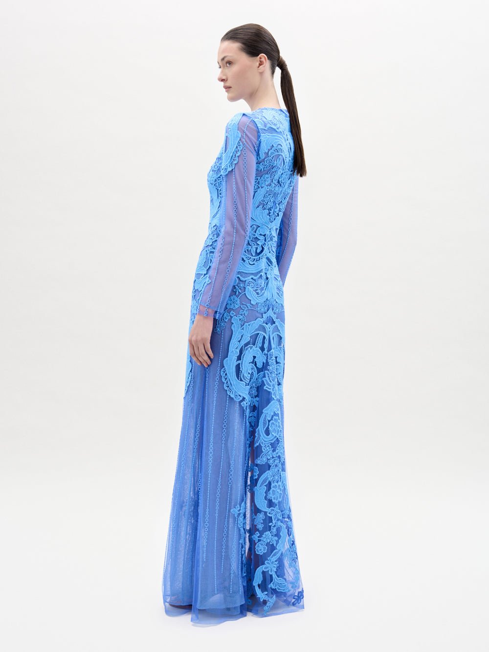 Una mujer mira hacia otro lado y lleva el vestido Lacey, de manga larga y largo hasta el suelo, de encaje azul, fabricado en Colombia. Lleva el pelo oscuro recogido en una coleta baja y posa ante un fondo sencillo y claro.