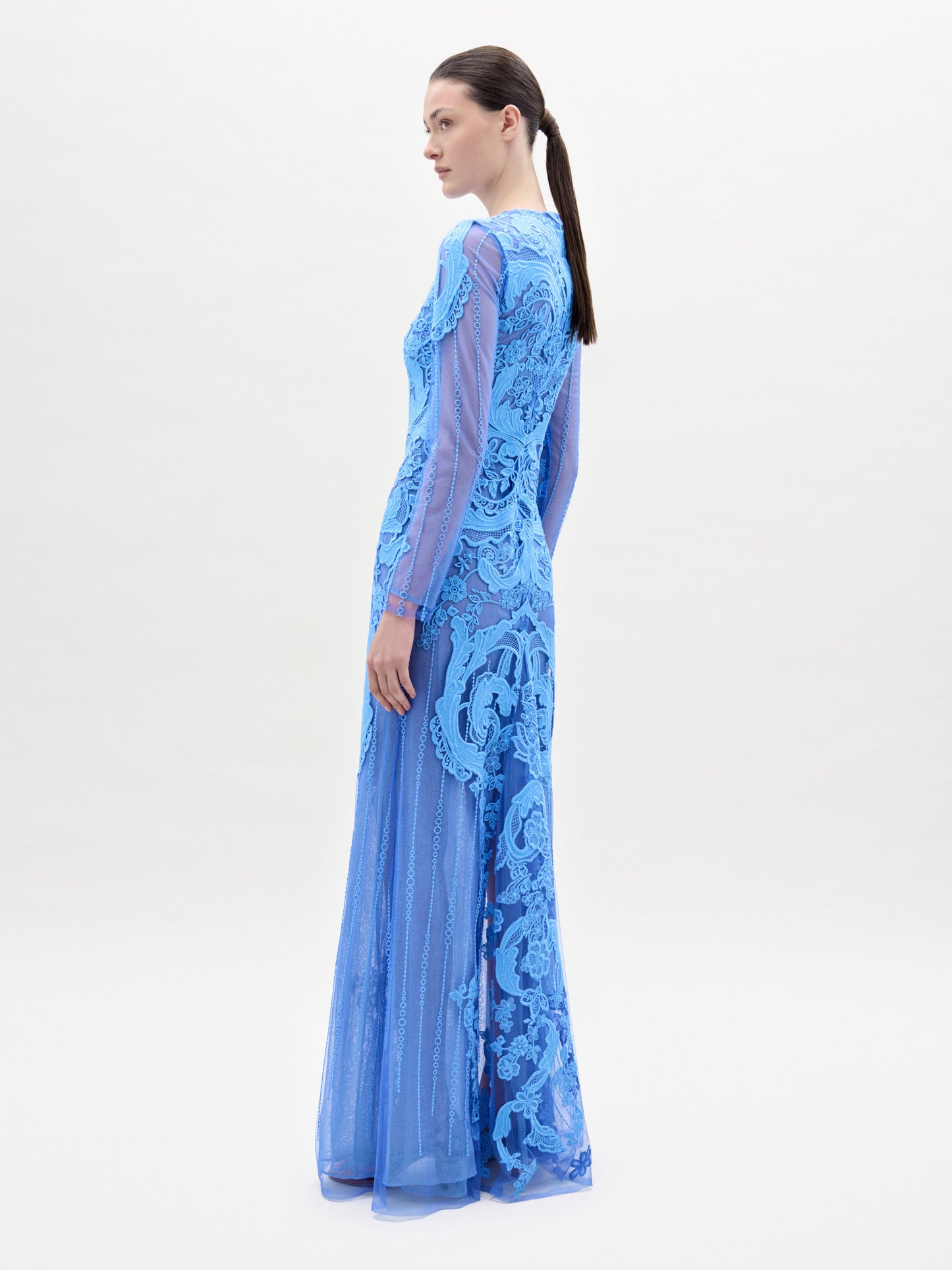 Una mujer mira hacia otro lado y lleva el vestido Lacey, de manga larga y largo hasta el suelo, de encaje azul, fabricado en Colombia. Lleva el pelo oscuro recogido en una coleta baja y posa ante un fondo sencillo y claro.