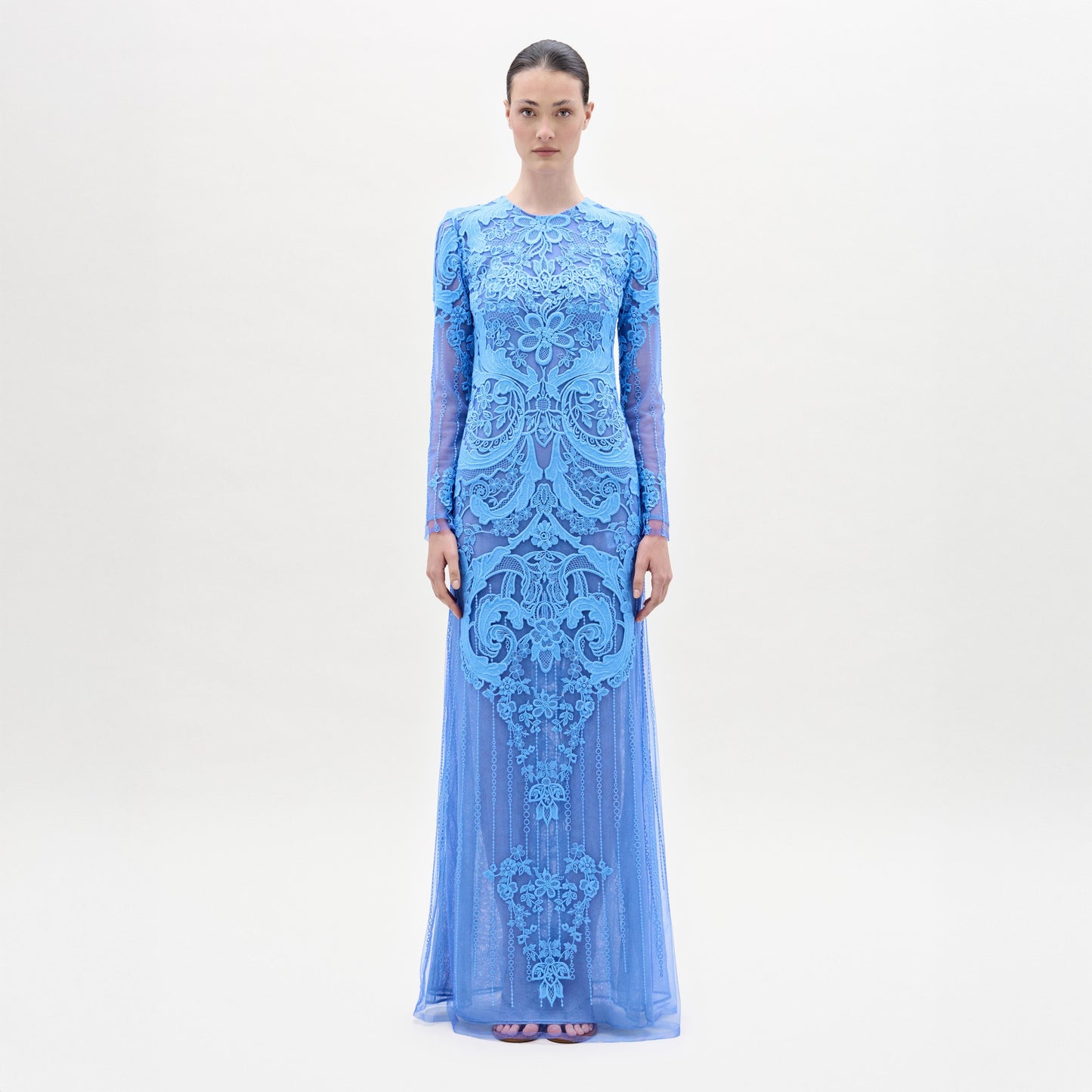 Una mujer de pie, de frente, lleva el vestido Lacey, largo y entallado, de color azul claro, con intrincados motivos florales de encaje y bordados, mangas largas, escote alto y bajo hasta el suelo, sobre un fondo blanco liso.