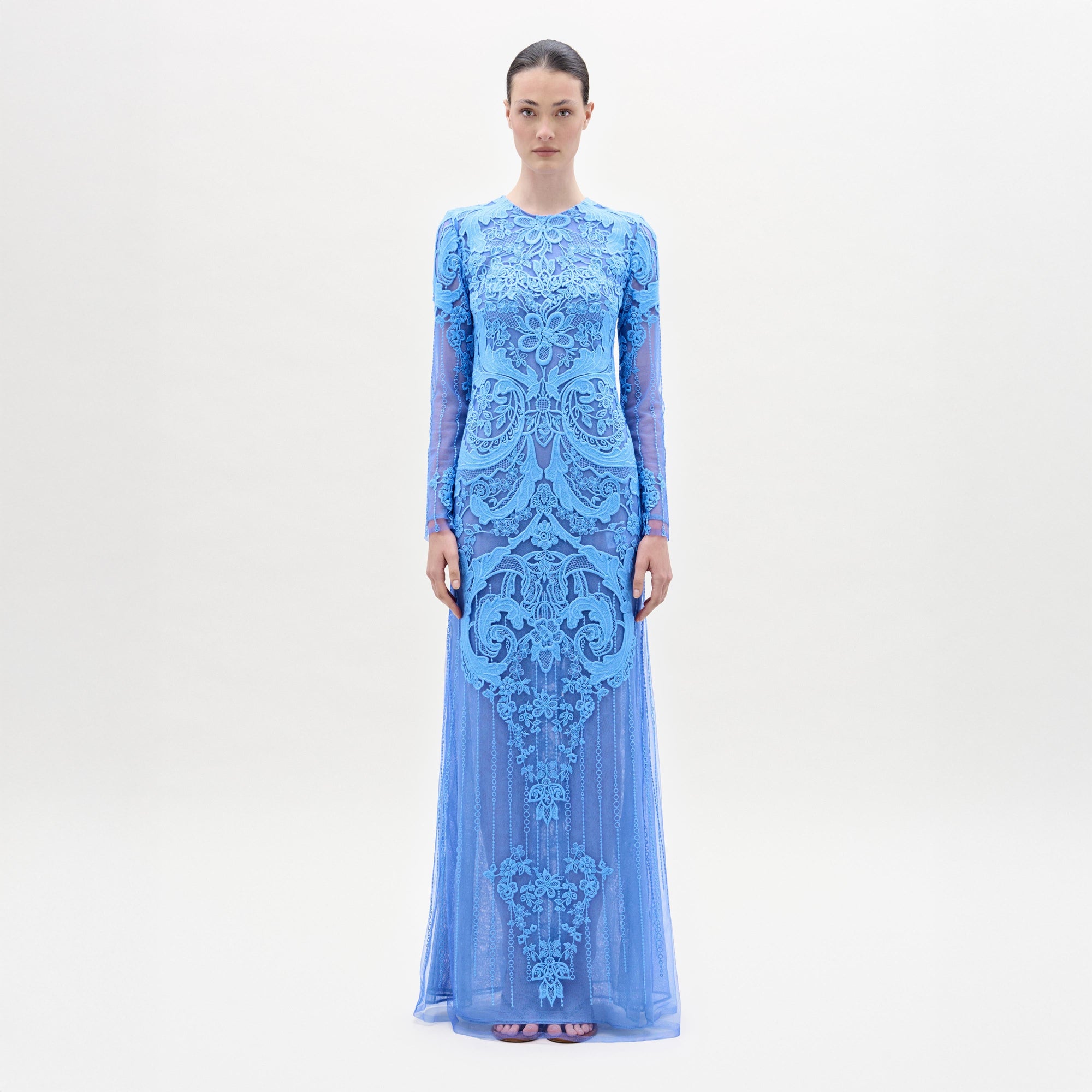 Una mujer de pie, de frente, lleva el vestido Lacey, largo y entallado, de color azul claro, con intrincados motivos florales de encaje y bordados, mangas largas, escote alto y bajo hasta el suelo, sobre un fondo blanco liso.