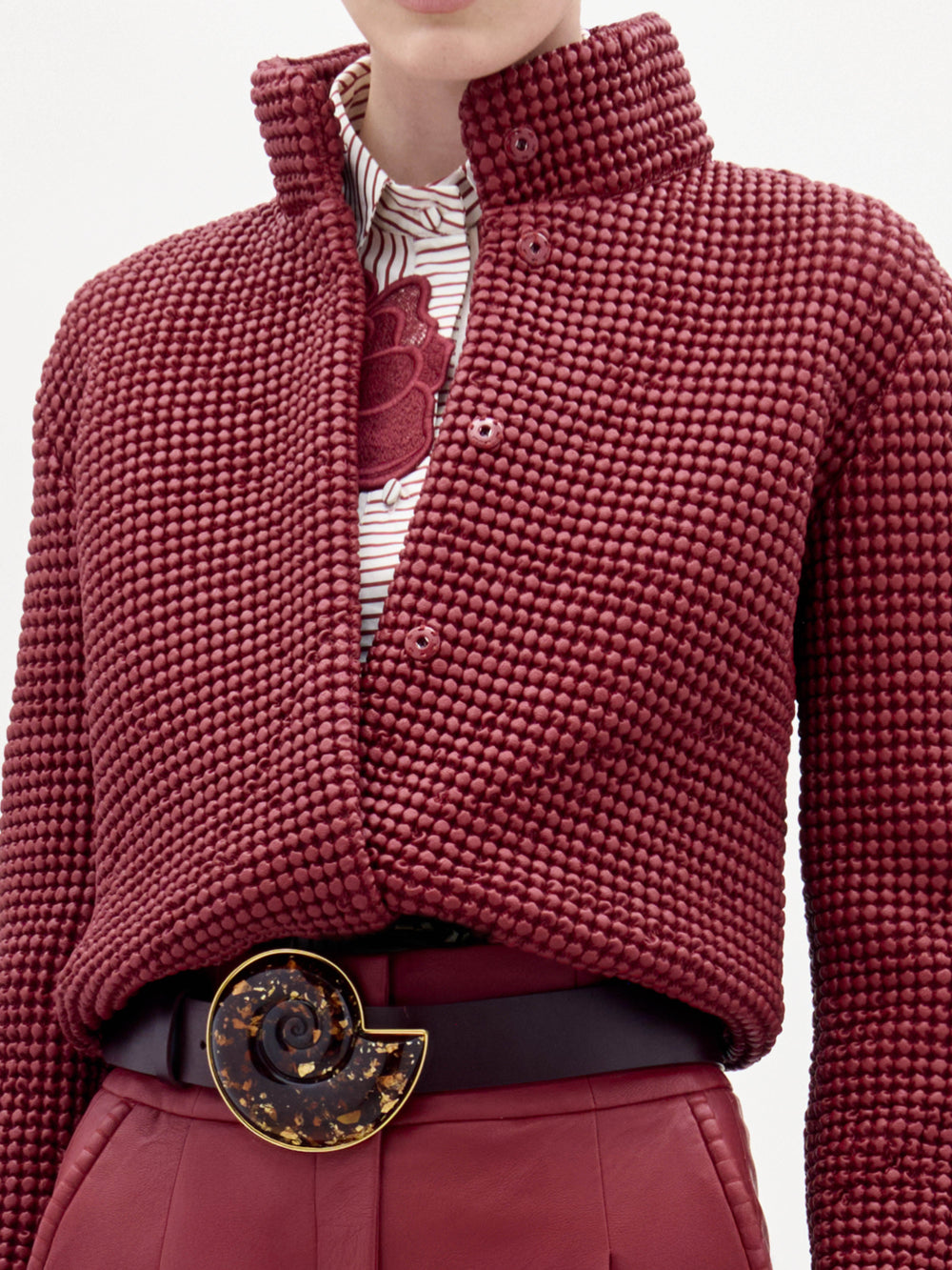 Una persona lleva la chaqueta Zalta -de color rojo oscuro, con textura y cuello alto- sobre una camisa blanca con estampado floral rojo, combinada con pantalones rojos a juego y un cinturón negro con una gran hebilla circular.