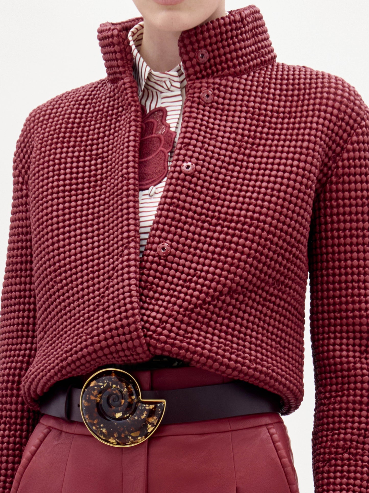 Una persona lleva la chaqueta Zalta -de color rojo oscuro, con textura y cuello alto- sobre una camisa blanca con estampado floral rojo, combinada con pantalones rojos a juego y un cinturón negro con una gran hebilla circular.