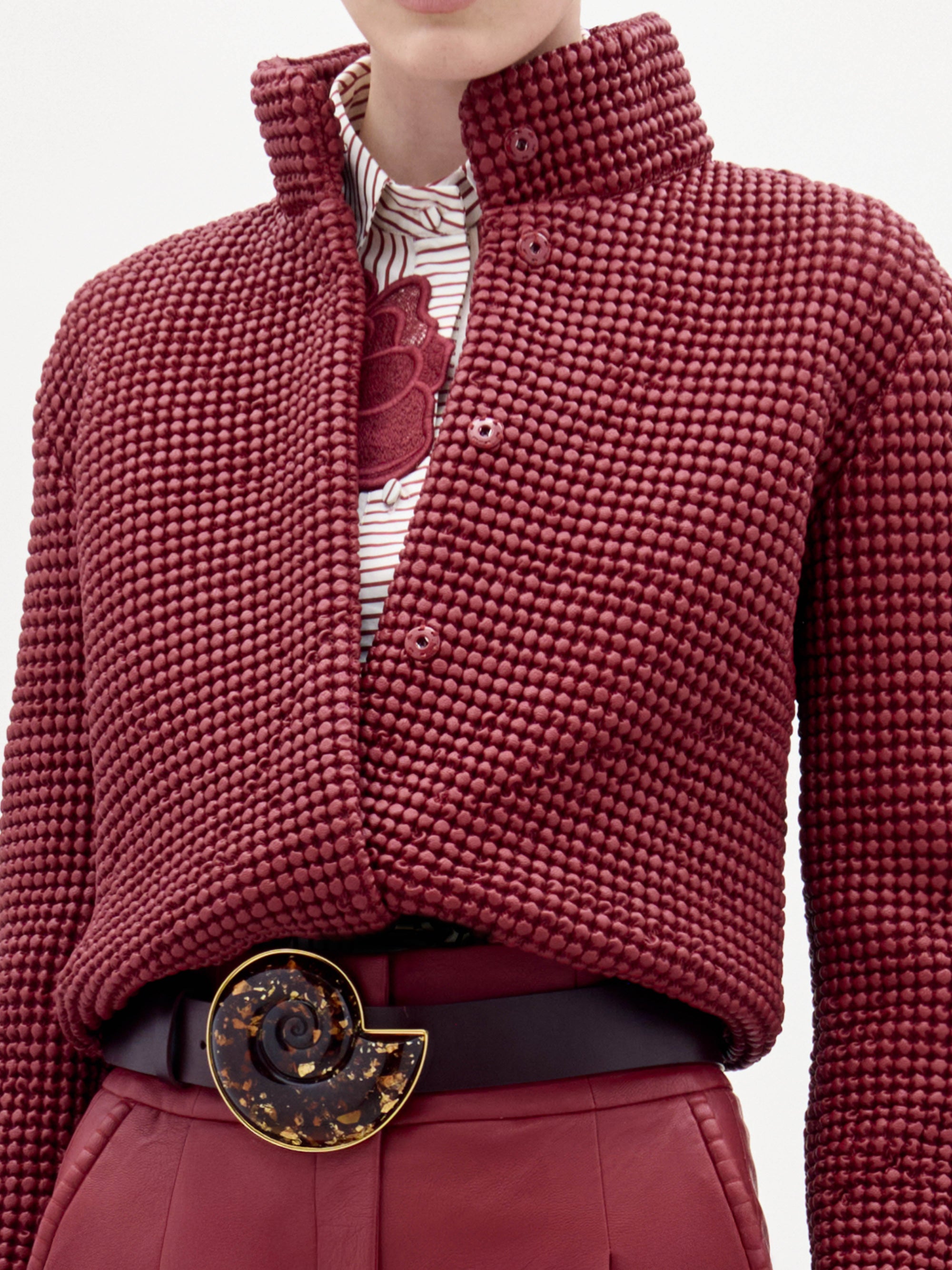 Una persona lleva la chaqueta Zalta -de color rojo oscuro, con textura y cuello alto- sobre una camisa blanca con estampado floral rojo, combinada con pantalones rojos a juego y un cinturón negro con una gran hebilla circular.