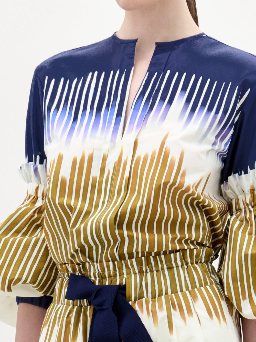 Una mujer lleva la blusa Lucaya, un top de manga larga en azul marino, blanco y dorado con escote dividido, mangas fruncidas, rayas verticales y un cinturón de lazo azul oscuro en la cintura. Confeccionada en tejido italiano de primera calidad.