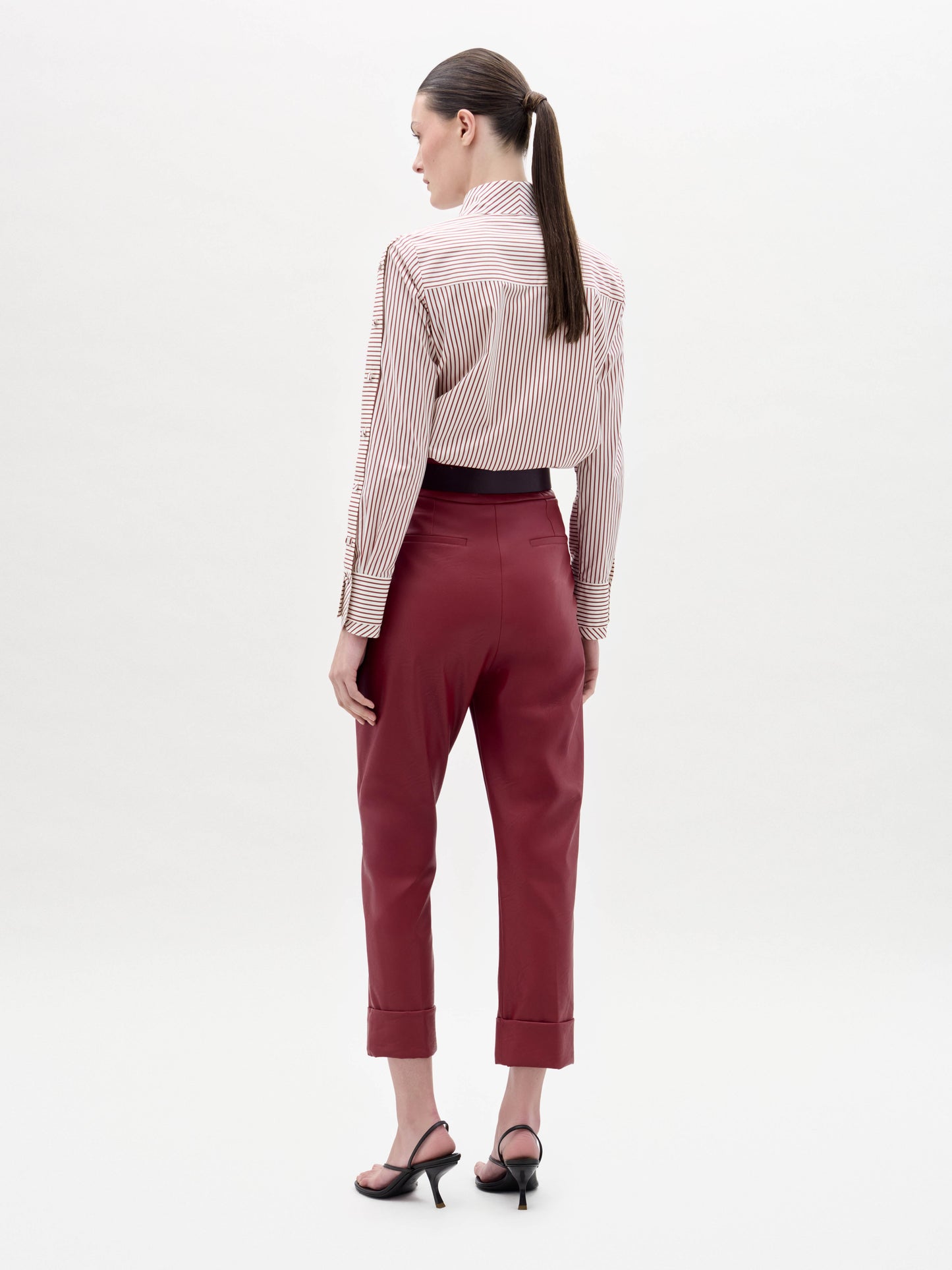 Una persona con el pelo largo recogido en una coleta mira hacia otro lado, llevando el Moad Pant -un pantalón rojo de cintura alta- con una camisa de rayas rojas y blancas y sandalias de tacón negras sobre un fondo liso de color claro.