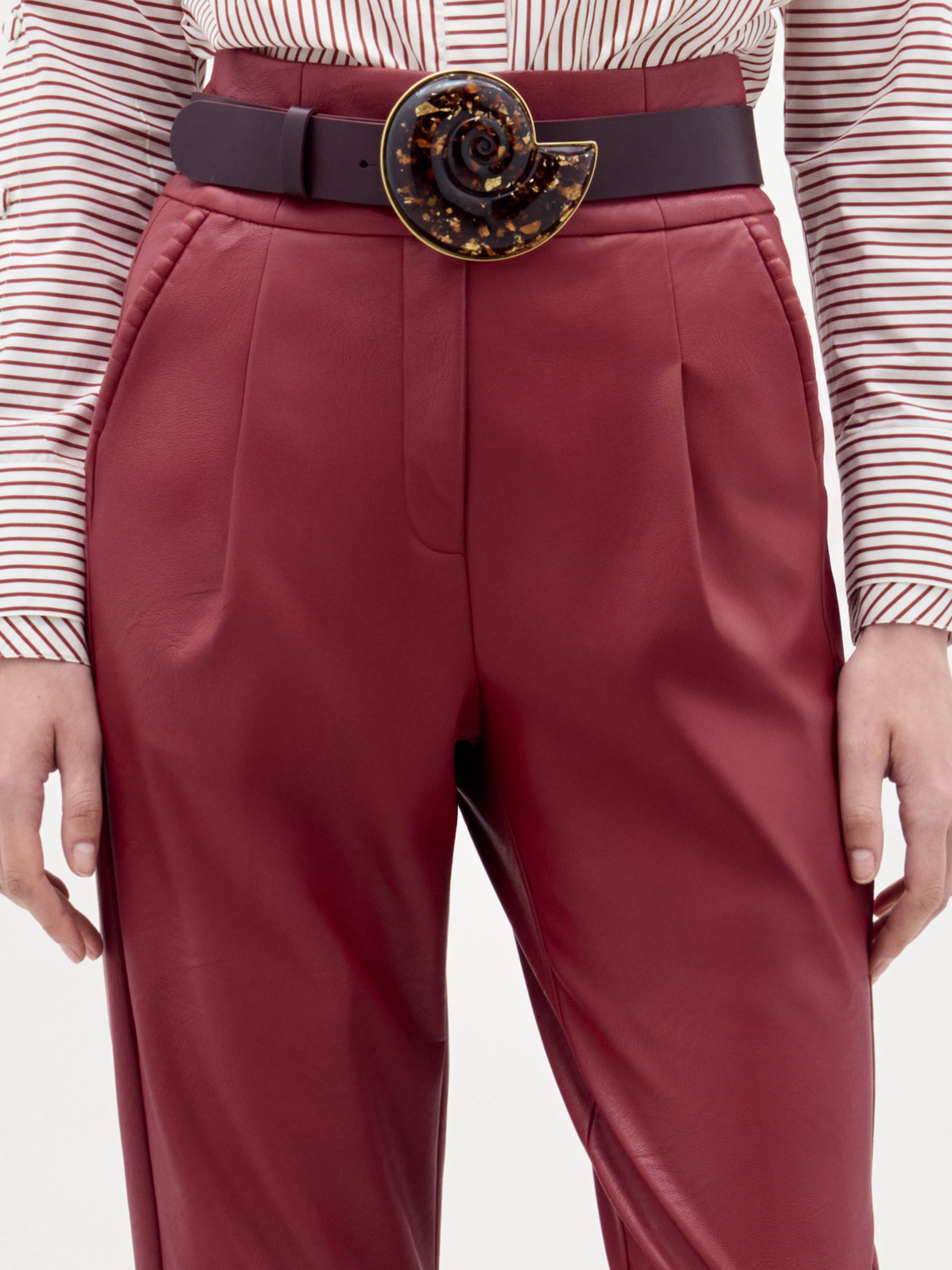 Una persona lleva el pantalón Moad -de talle alto y color burdeos- con una camisa de manga larga a rayas y un cinturón ancho con una gran hebilla redonda en forma de espiral. Sólo se ven el torso y la parte superior de las piernas.