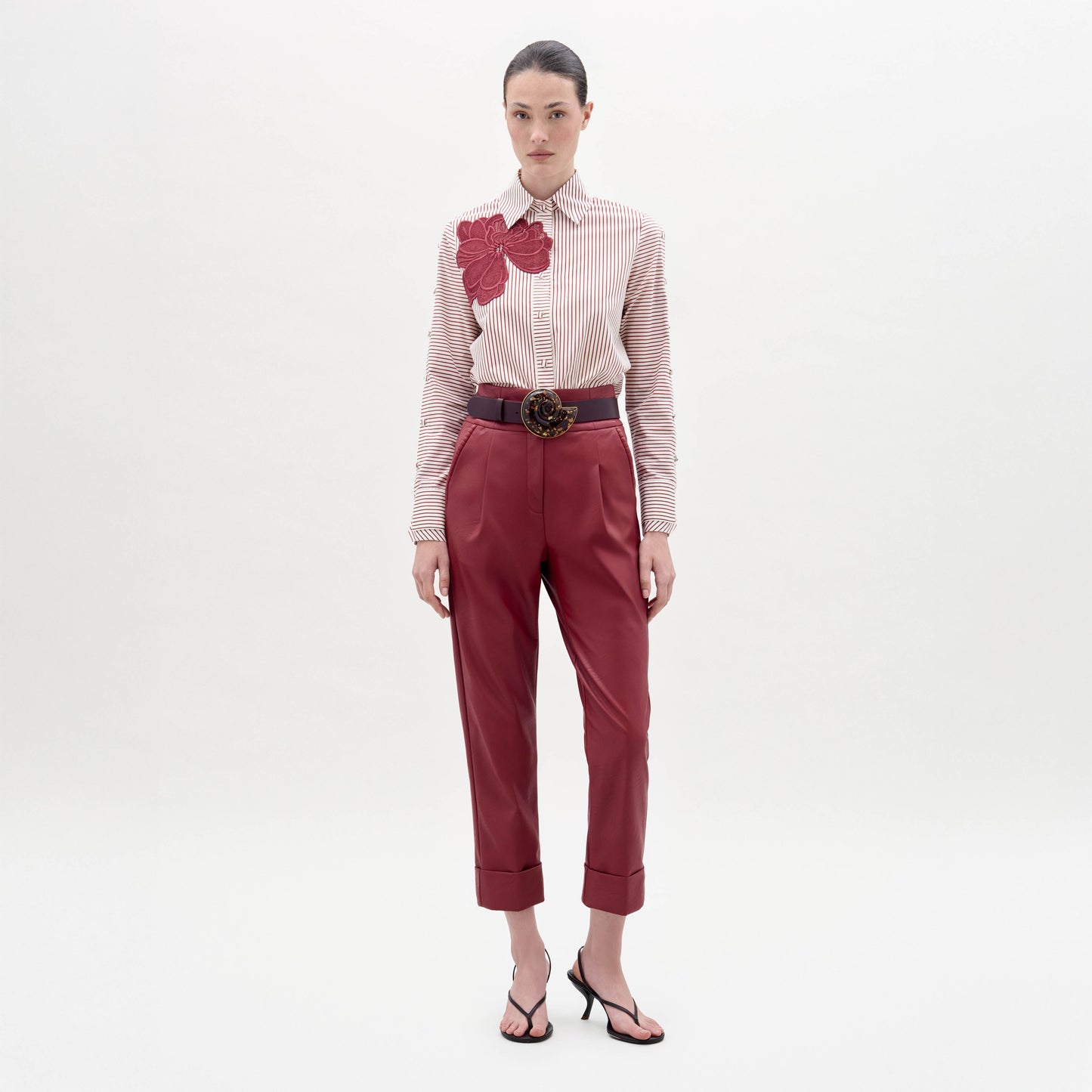 Una mujer posa sobre un fondo blanco liso con el pantalón Moad Pant -pantalón rojo de cintura alta con puños- combinado con una camisa de rayas con un gran aplique floral rojo, un cinturón ancho con hebilla decorativa y sandalias negras.