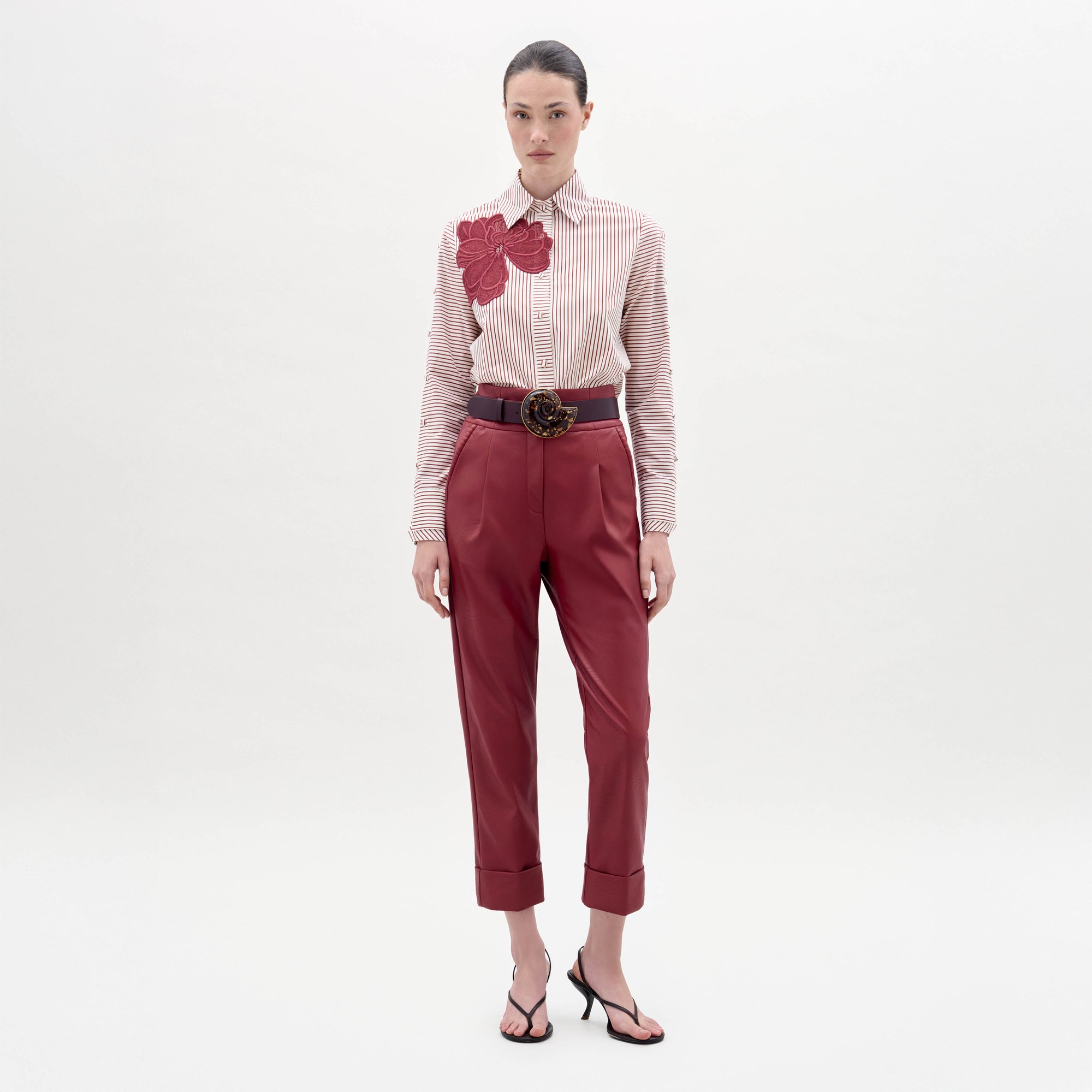 Una mujer posa sobre un fondo blanco liso con el pantalón Moad Pant -pantalón rojo de cintura alta con puños- combinado con una camisa de rayas con un gran aplique floral rojo, un cinturón ancho con hebilla decorativa y sandalias negras.