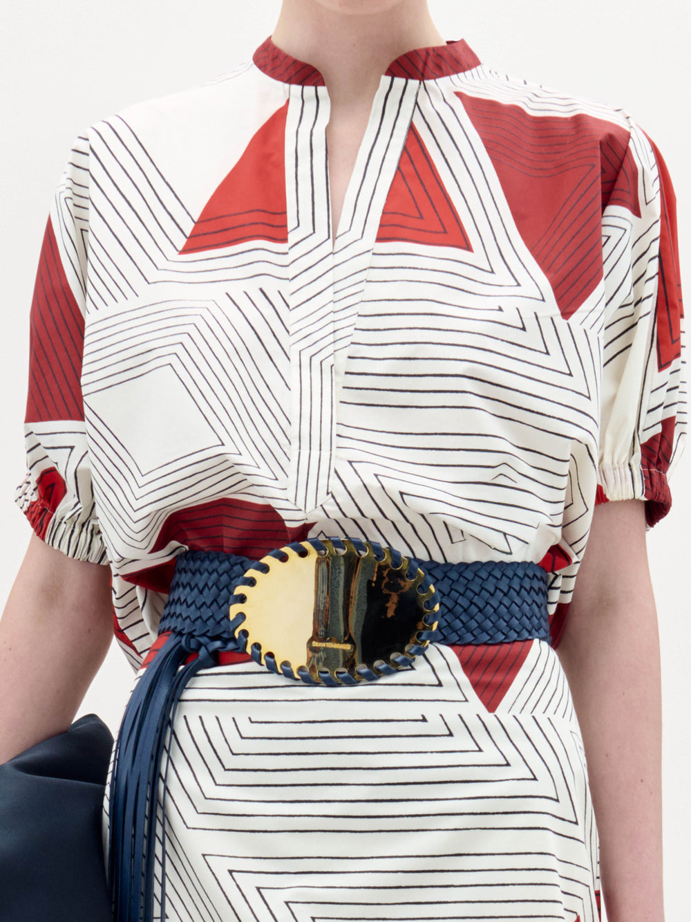 Una persona lleva la blusa Susanne -una blusa blanca con motivos geométricos rojos y finas líneas negras- combinada con un cinturón tejido azul con una gran hebilla metálica ovalada, y sostiene un bolso azul oscuro.